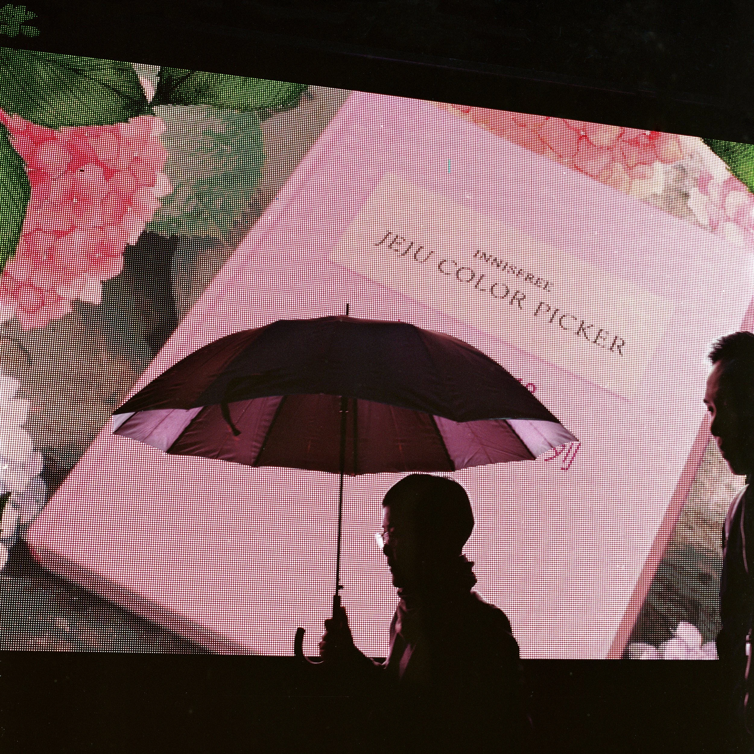 People walking with an umbrella in front of a large pink-colored advertisement featuring a box of pink color pencils with the text 'MINI PENCIL' and 'PRO COLOR PICKER' behind it.