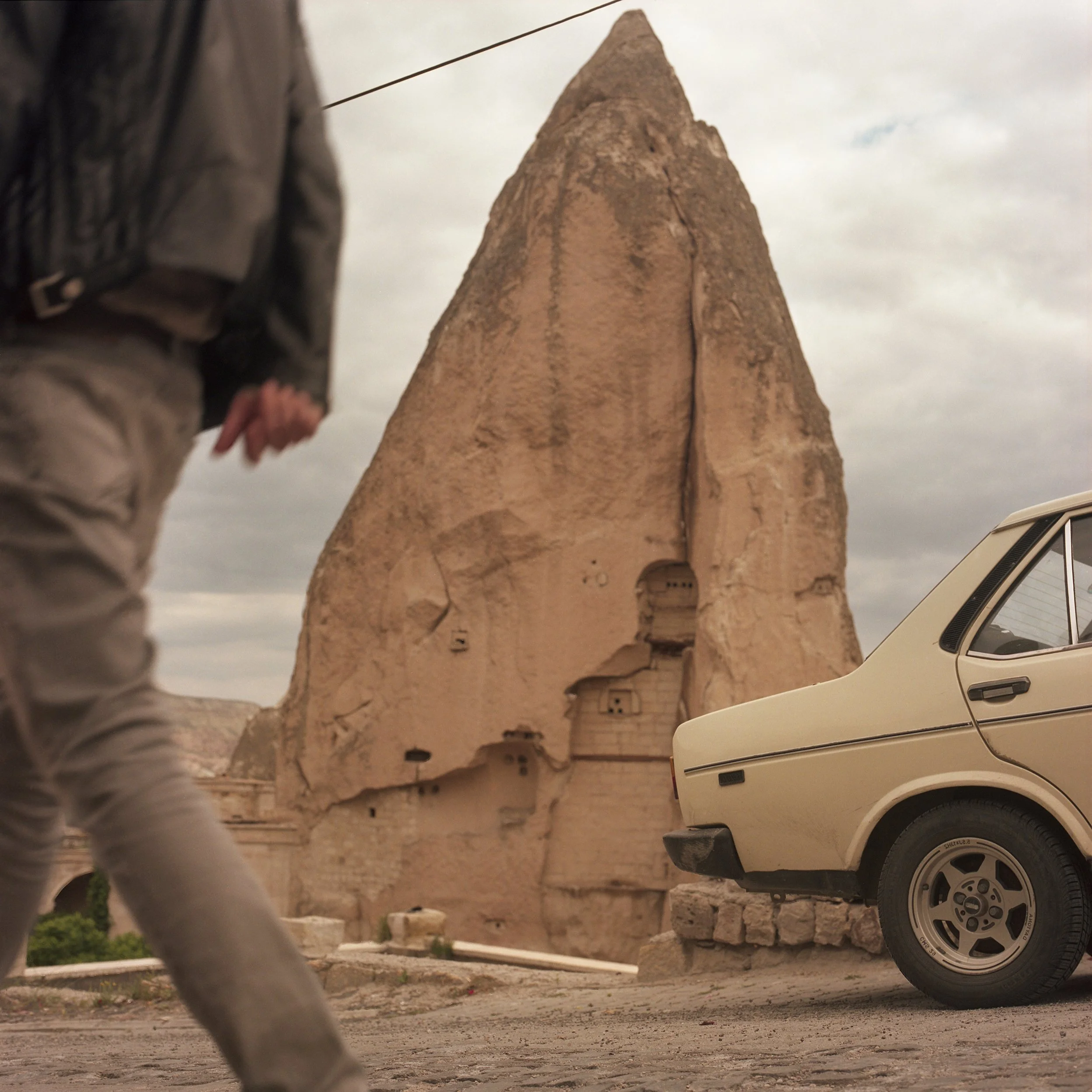 In Goreme, Turkey, A large rock formation with ancient caves carved into it, a vintage cream-colored car parked nearby, and a person walking on the street, under a cloudy sky.