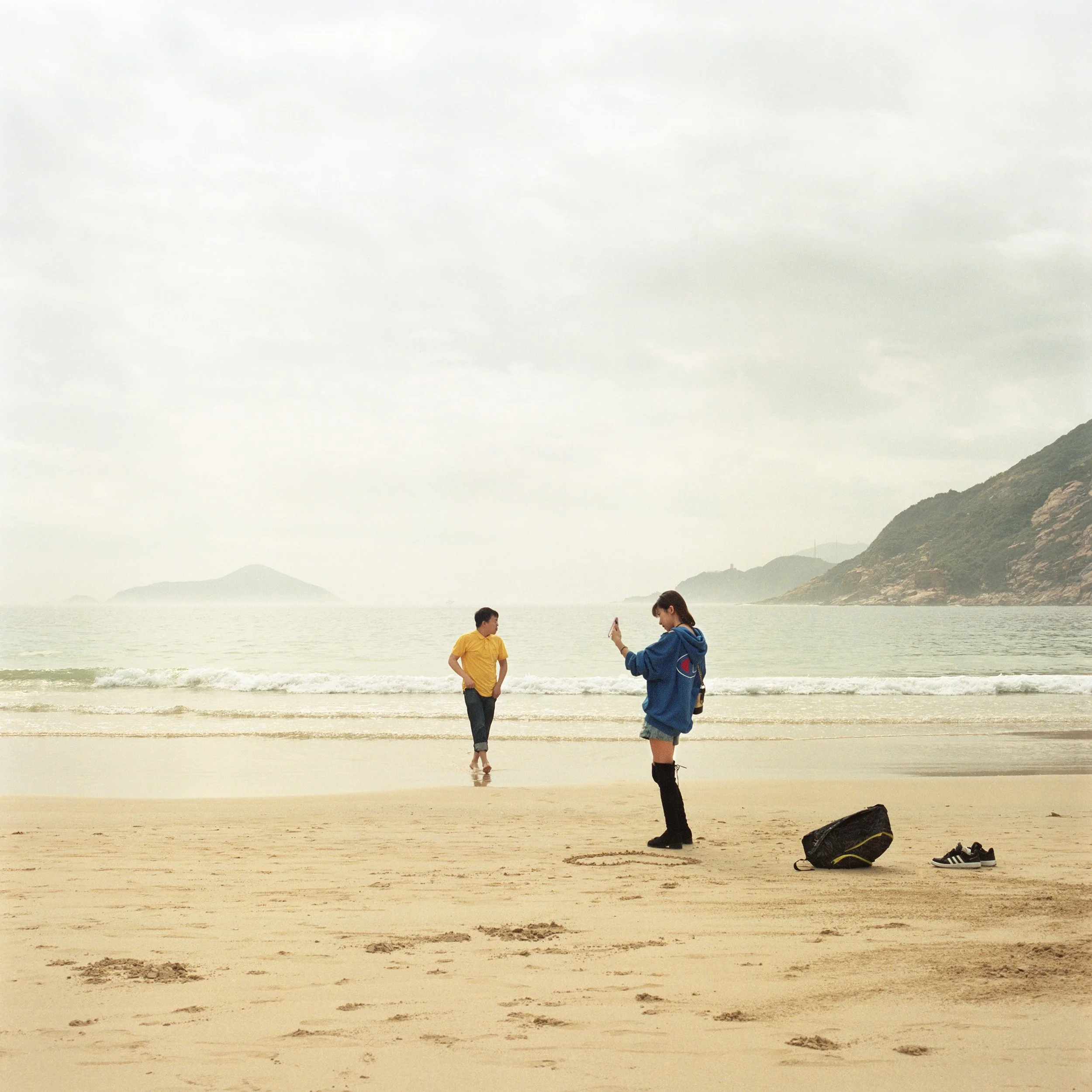 A person in a blue hoodie and shorts takes a photo on a cellphone at the beach, while a man in a yellow shirt stands near the shoreline with the ocean, small waves, islands, and mountains in the background.