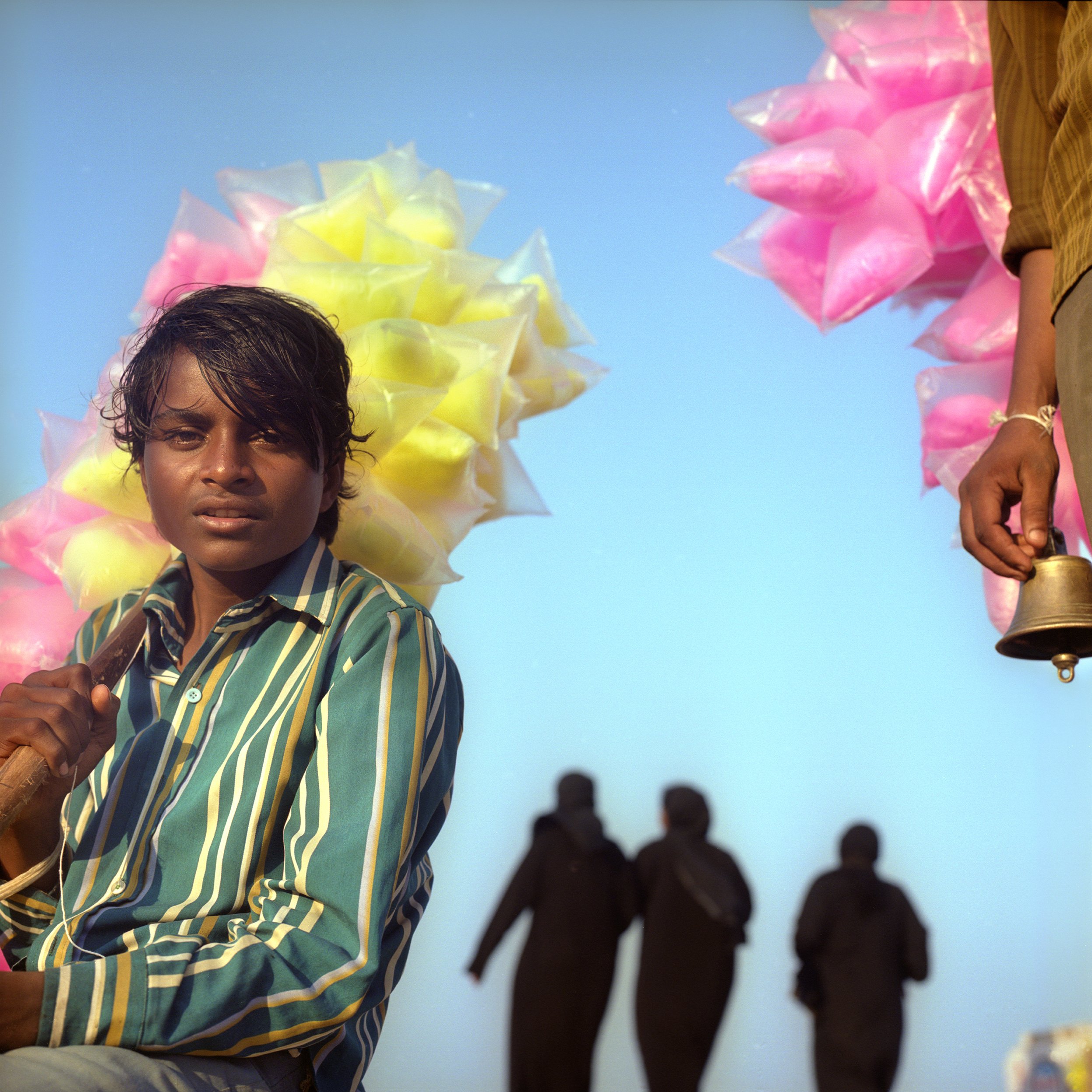 A young boy holding a stick with yellow and pink cotton candy, with colorful cotton candy in the background. Silhouettes of people walking in the distance against a clear blue sky.