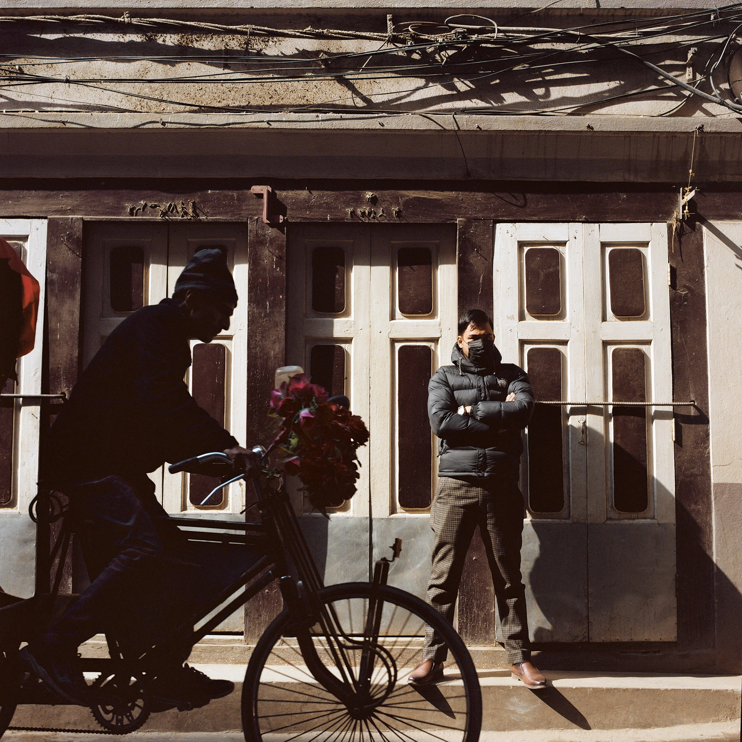 In Katmandu, A person on a bicycle carrying a bouquet of flowers passes a woman standing against a door, both wearing masks and warm clothing, with overhead wires visible.