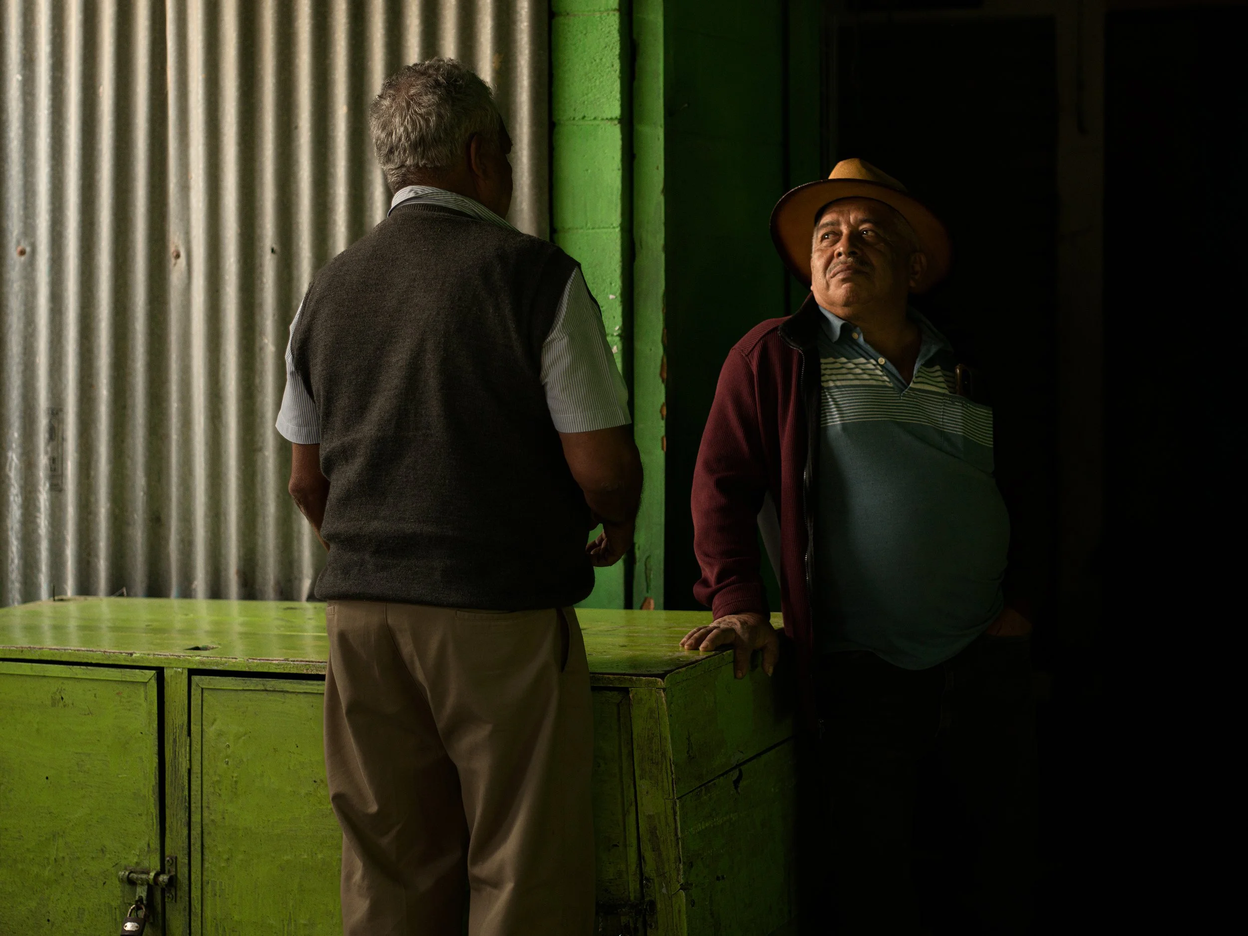 In Antigua market, Guatemala, Two men are engaged in conversation inside a dimly lit room, one leaning on a green wooden counter, both facing each other.