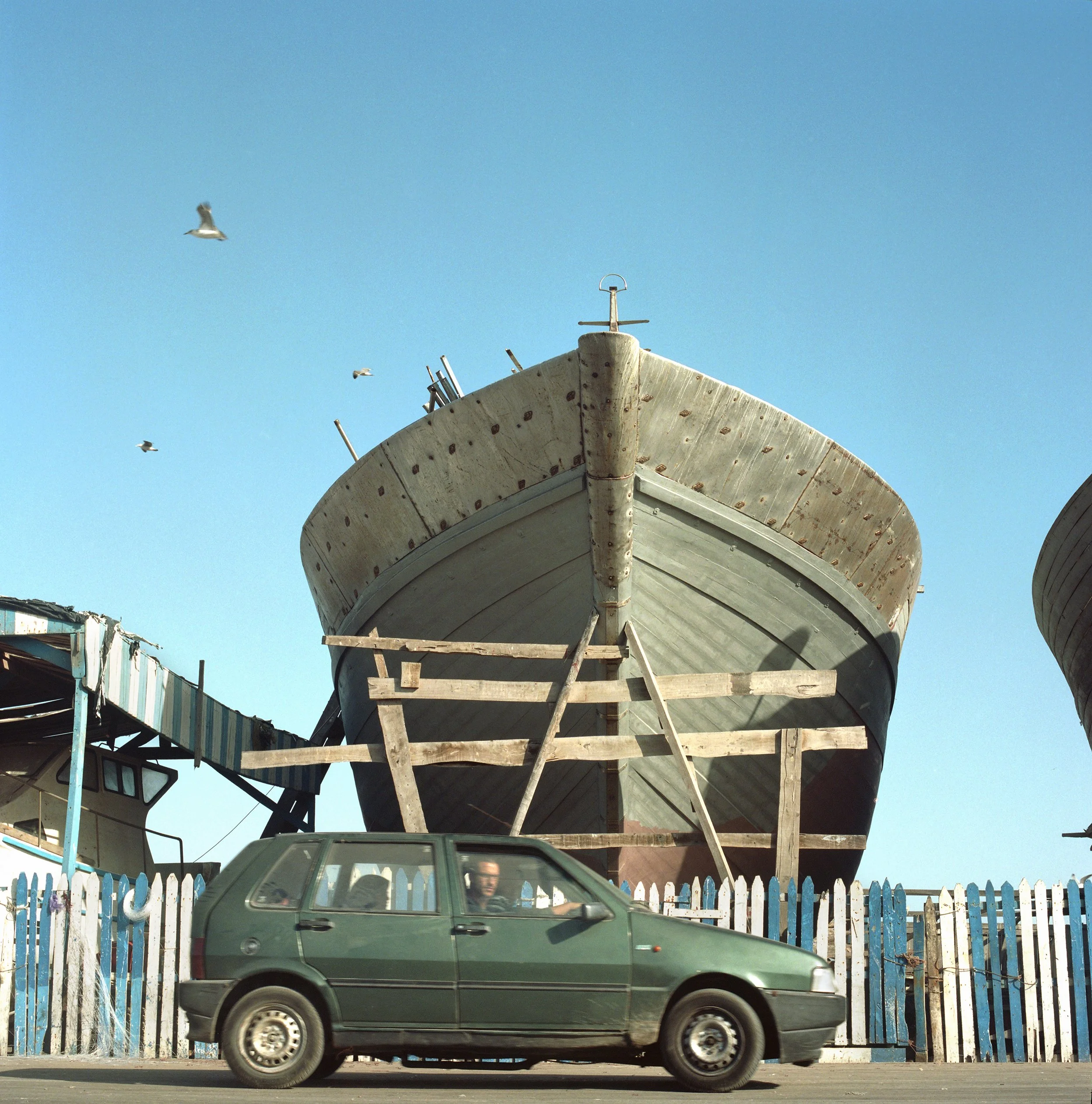 A boat on wooden supports behind a white fence, with a green car passing in front and seagulls flying in the blue sky.