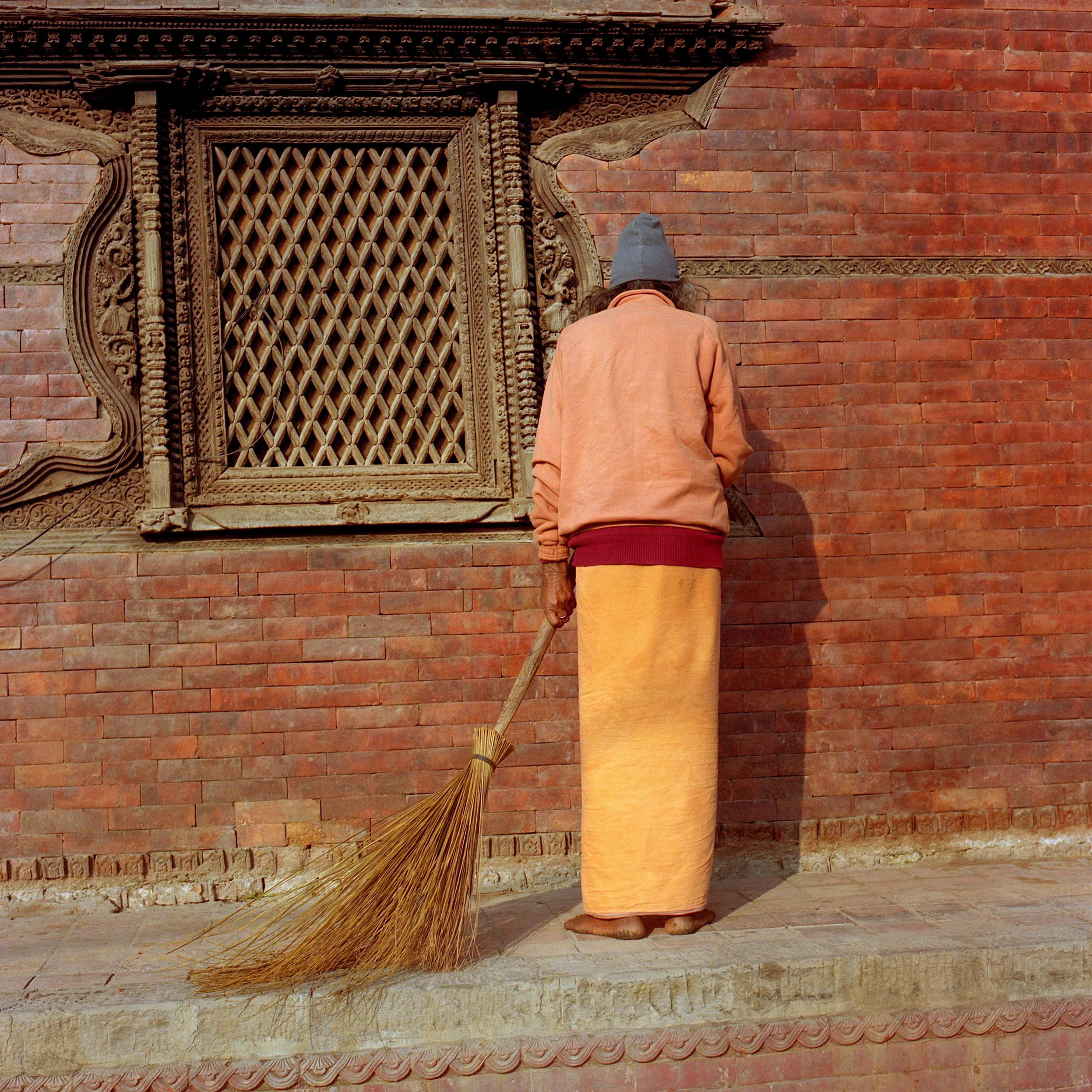 In Kathmandu, A person in traditional clothing sweeping the ground with a broom in front of a red brick wall with decorative wooden window frame.