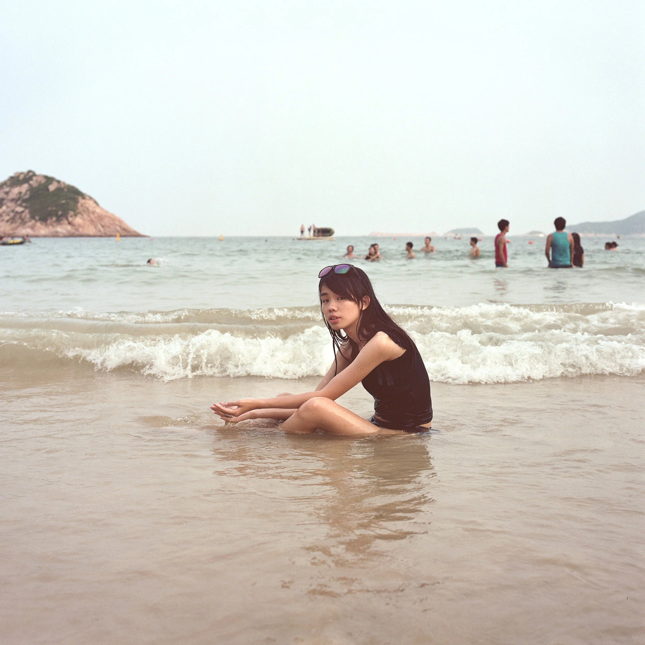 Young girl in black swimsuit sitting in shallow water at the beach with others swimming and standing in the background, and a small island on the horizon.