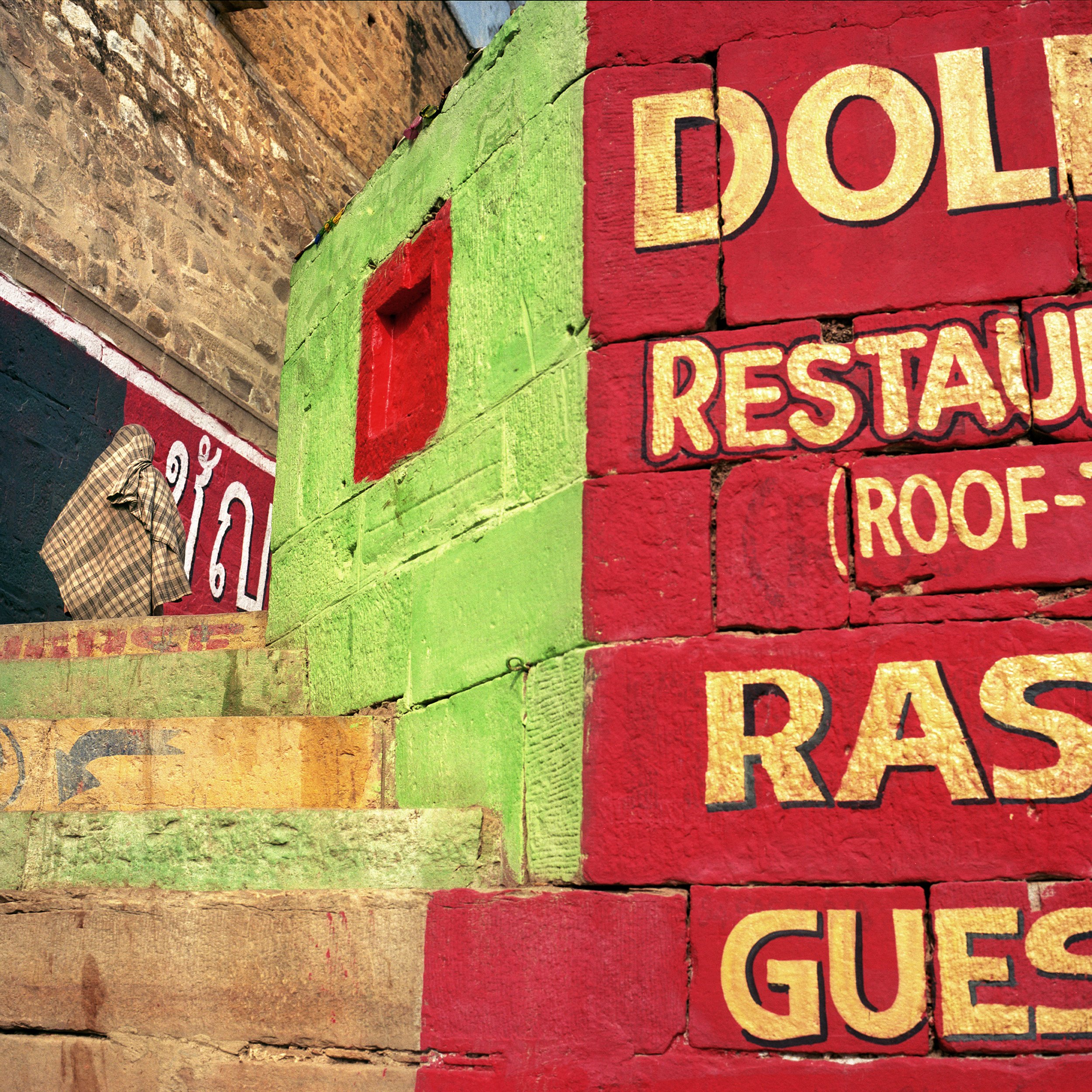 Close-up of a colorful brick wall with painted text and a small red window. The wall features red, green, and yellow brick sections, with some bricks showing signs of wear and cracks.