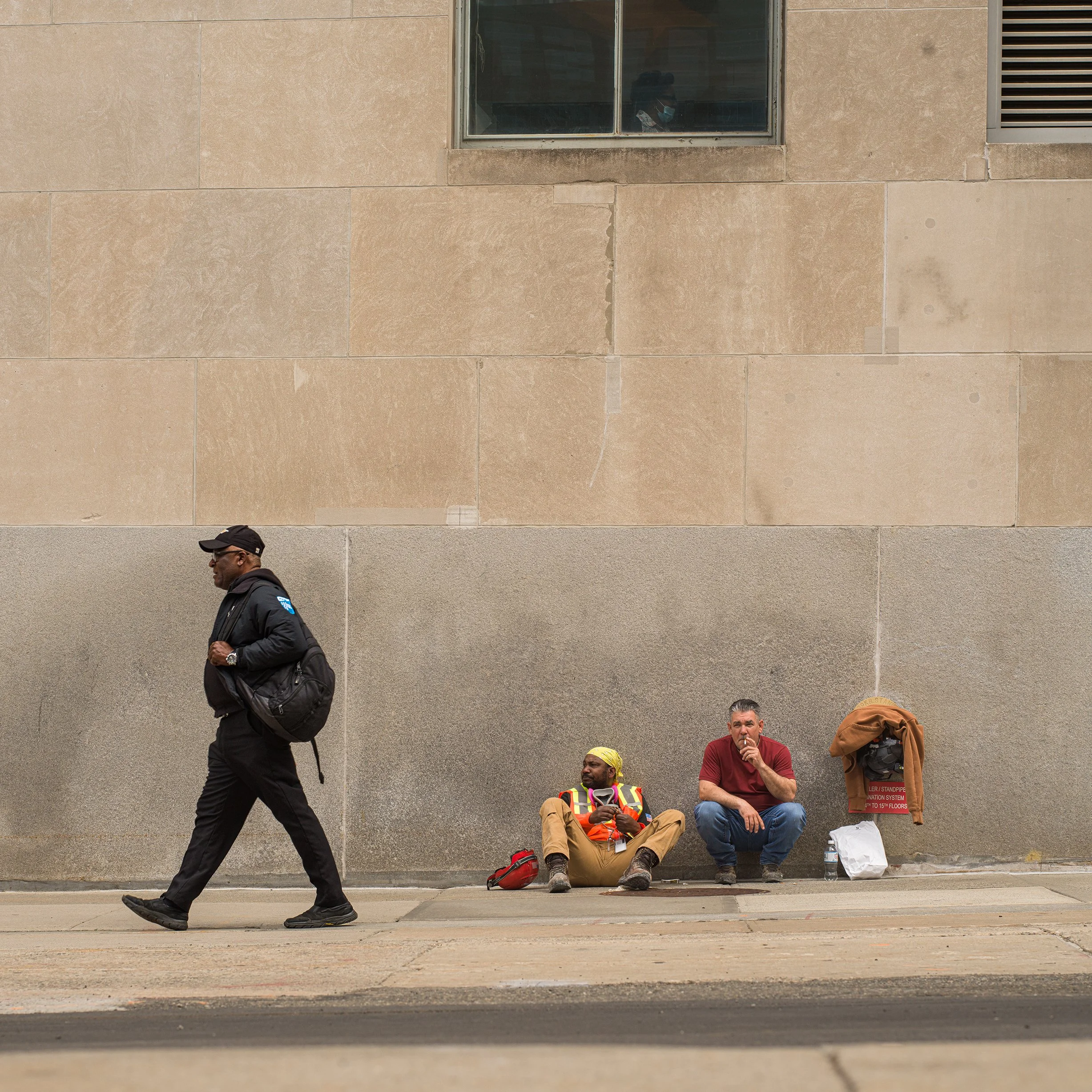 	
In New York Manhattan, A man walking past two people sitting on the sidewalk against a large beige wall, with a window above them. 