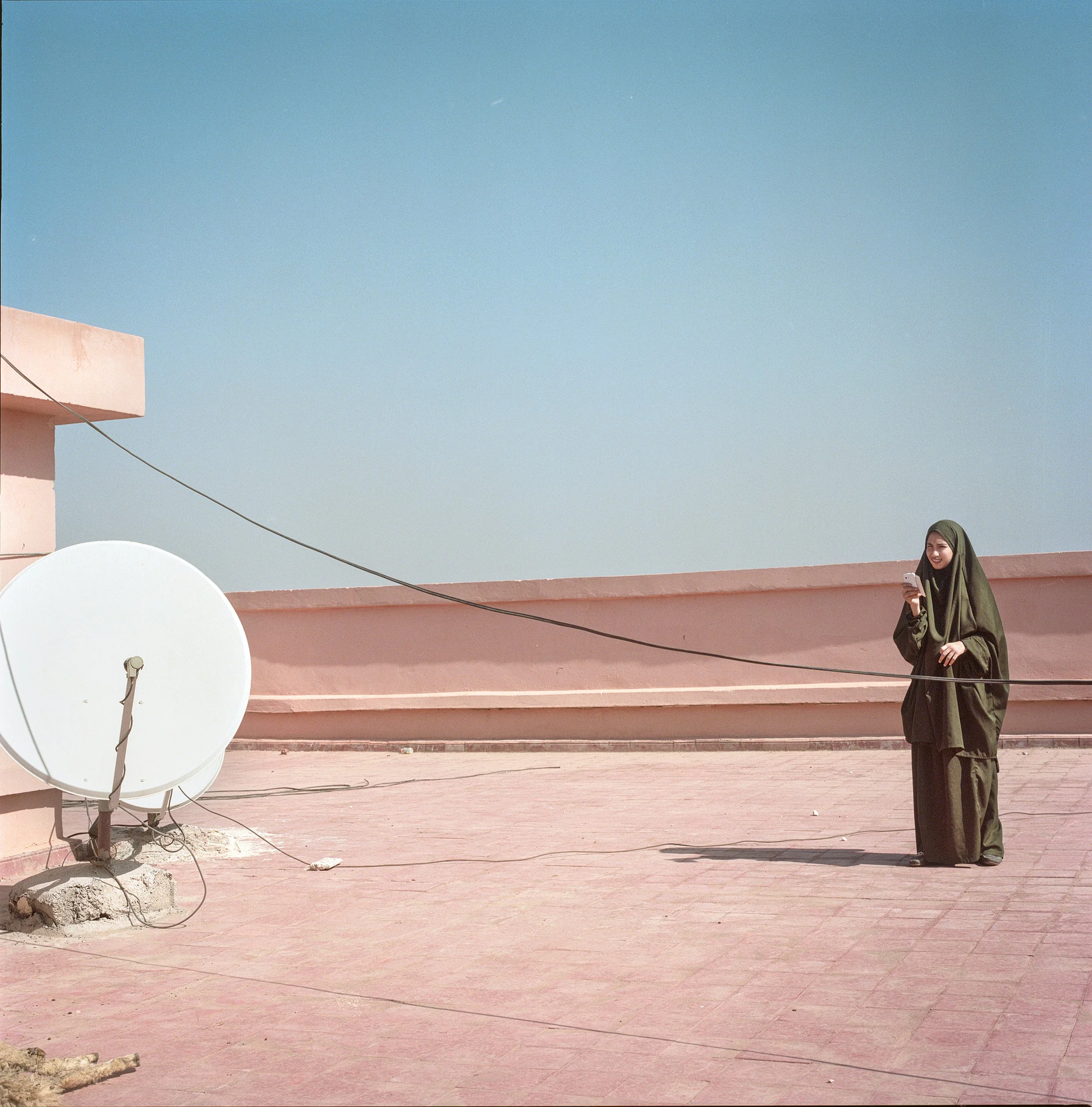 A woman wearing a dark green hijab and dress, standing on a pink rooftop, looking at her phone and smiling with a clear blue sky in the background.