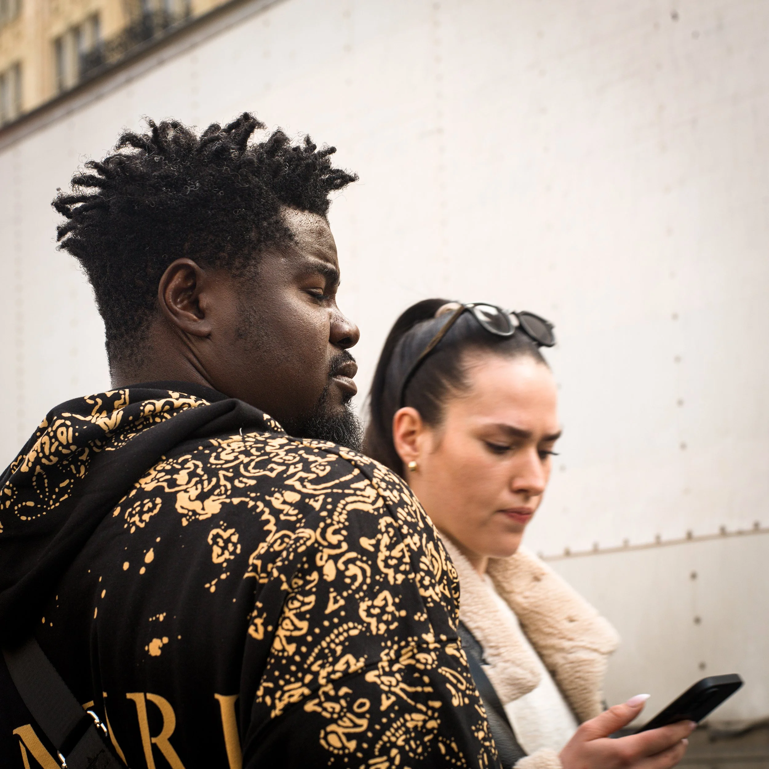 In New York Manhattan, A man wearing a black and tan patterned hoodie looks down with a serious expression, standing next to a woman with dark sunglasses on her head, and a beige furry coat, who is looking at her phone.