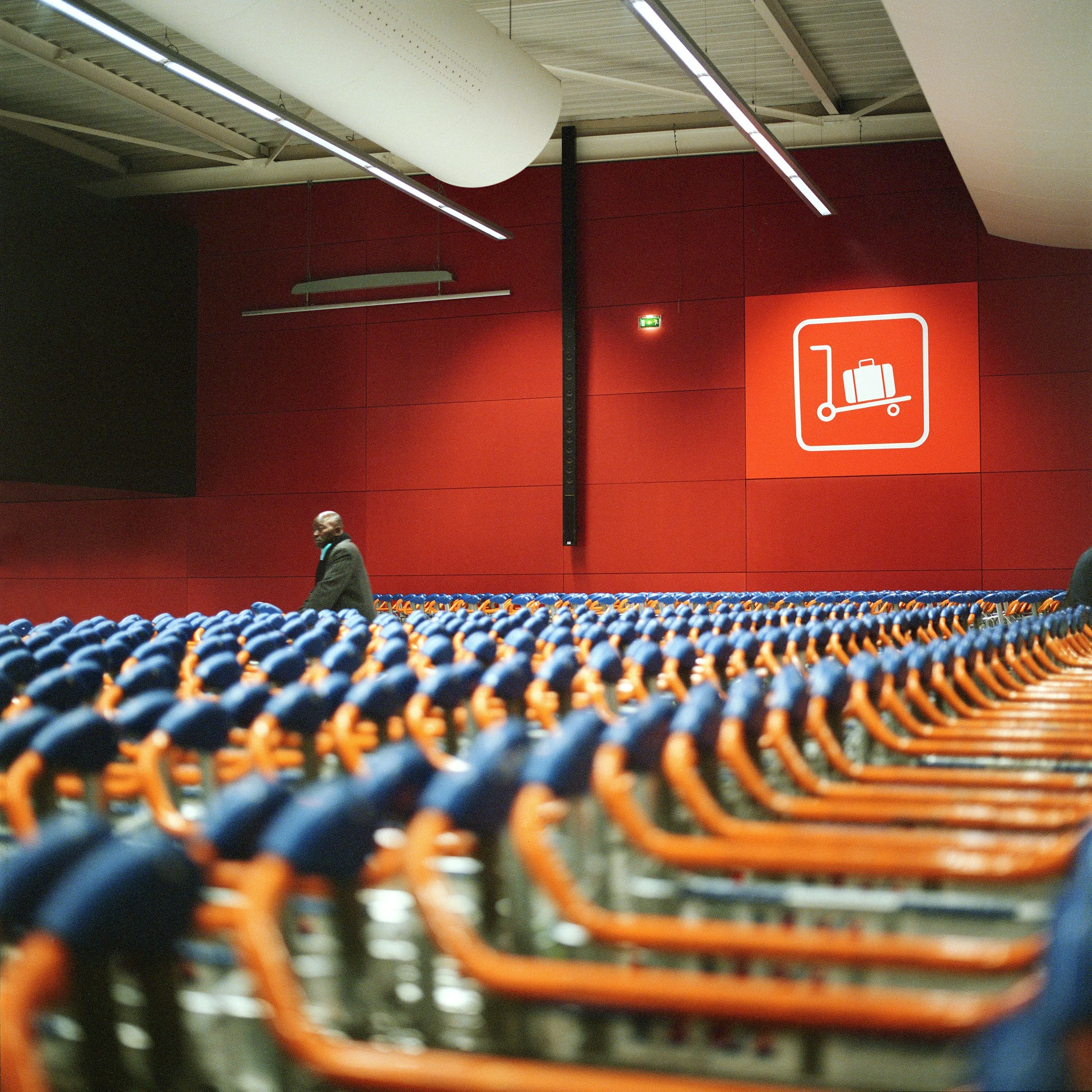 Rows of orange shopping carts in an airport terminal with a red wall and a sign showing luggage on a trolley.