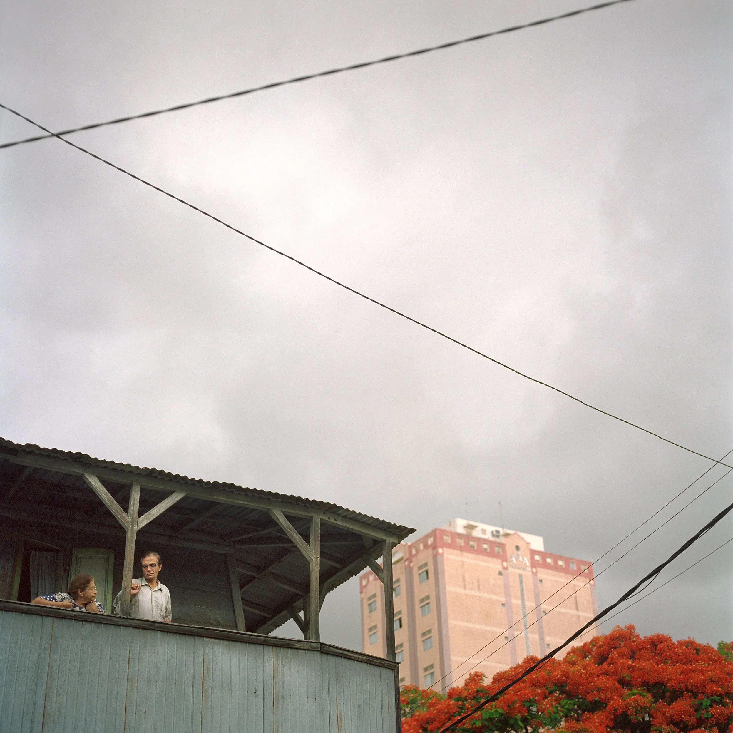 In Mauritius, Port-Louis, An overcast sky with utility cables, a wooden house with two people on the balcony, a tall pinkish building, and orange flowering trees.