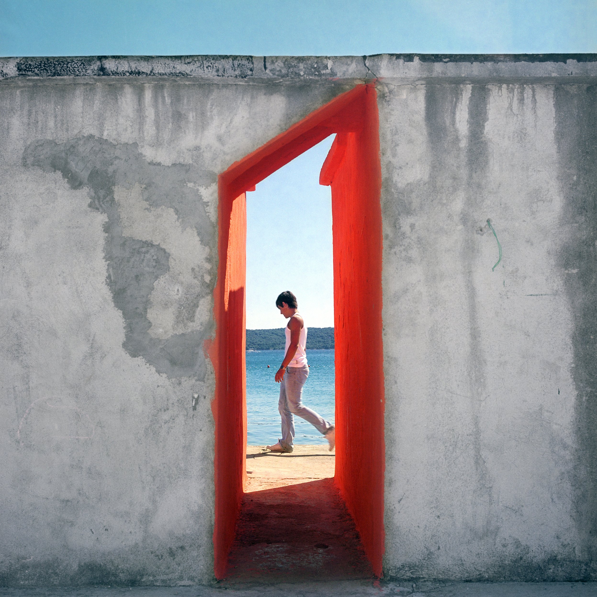 A person walks by a small opening in a concrete wall, with the beach and water visible through the opening.