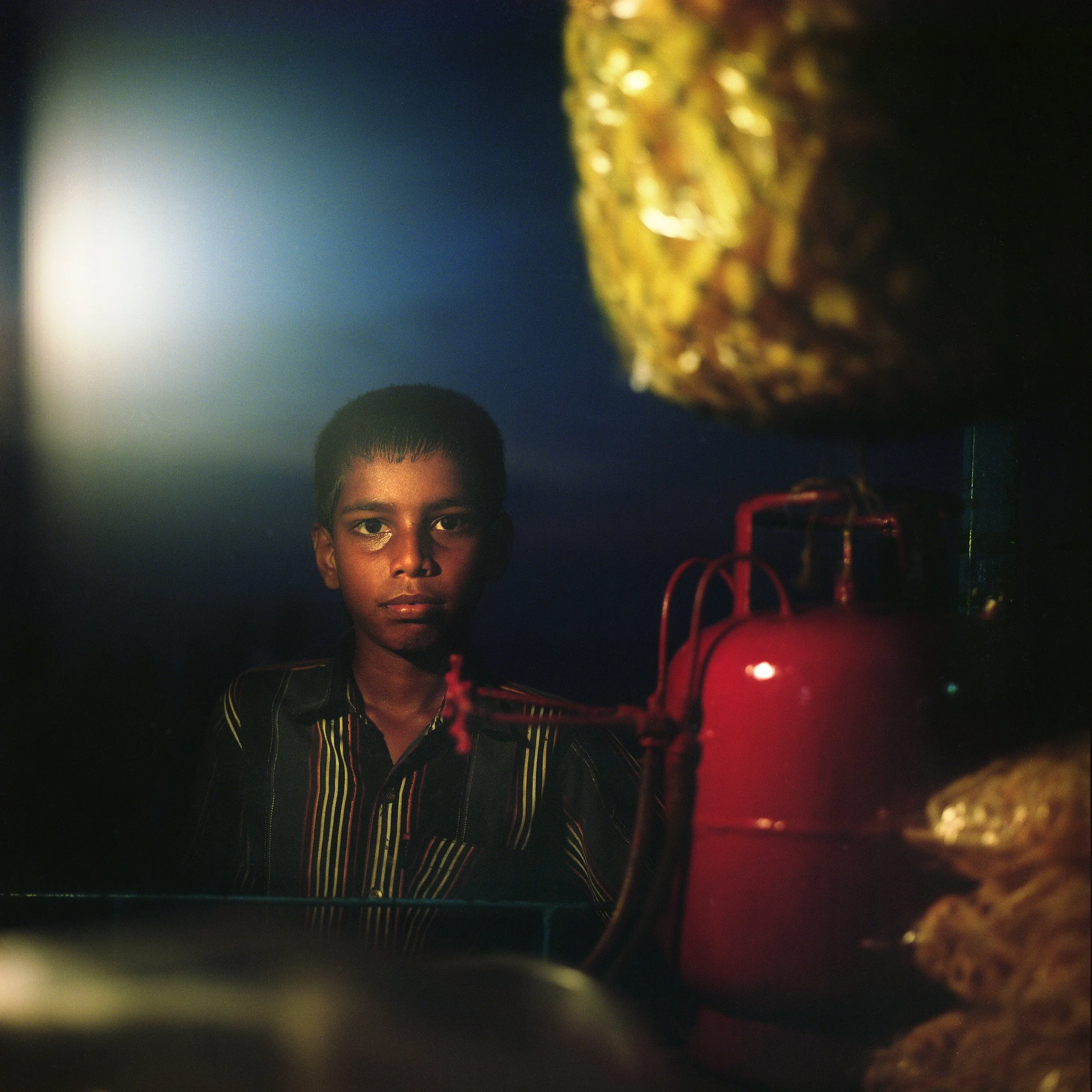 A young boy with dark skin and short hair looking through a refrigerator. The image is taken from inside the fridge, showing groceries and a red thermos in the foreground, with a dark blue background.