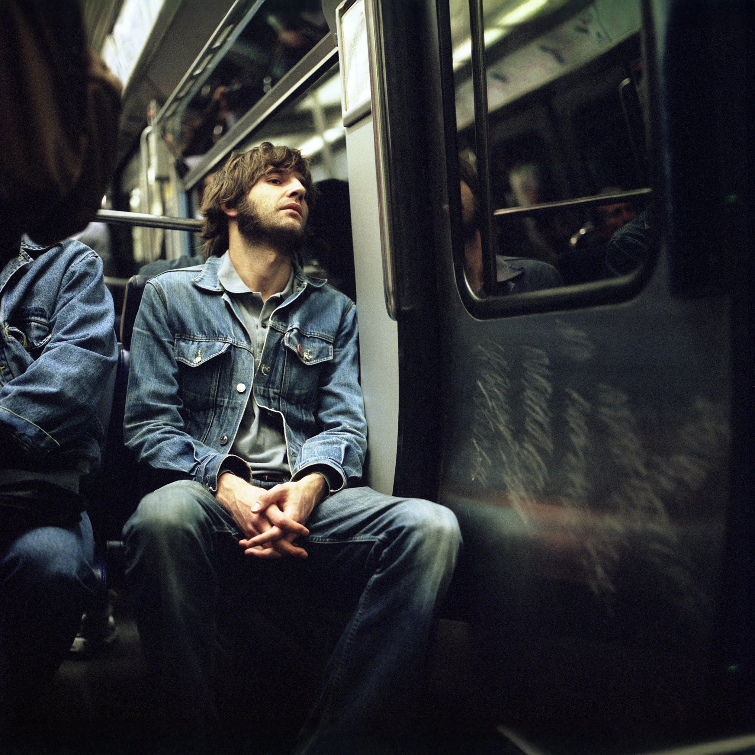 A young man with brown hair and a beard, wearing a denim jacket and grey shirt, is seated on a subway train, looking to the side with a neutral expression.