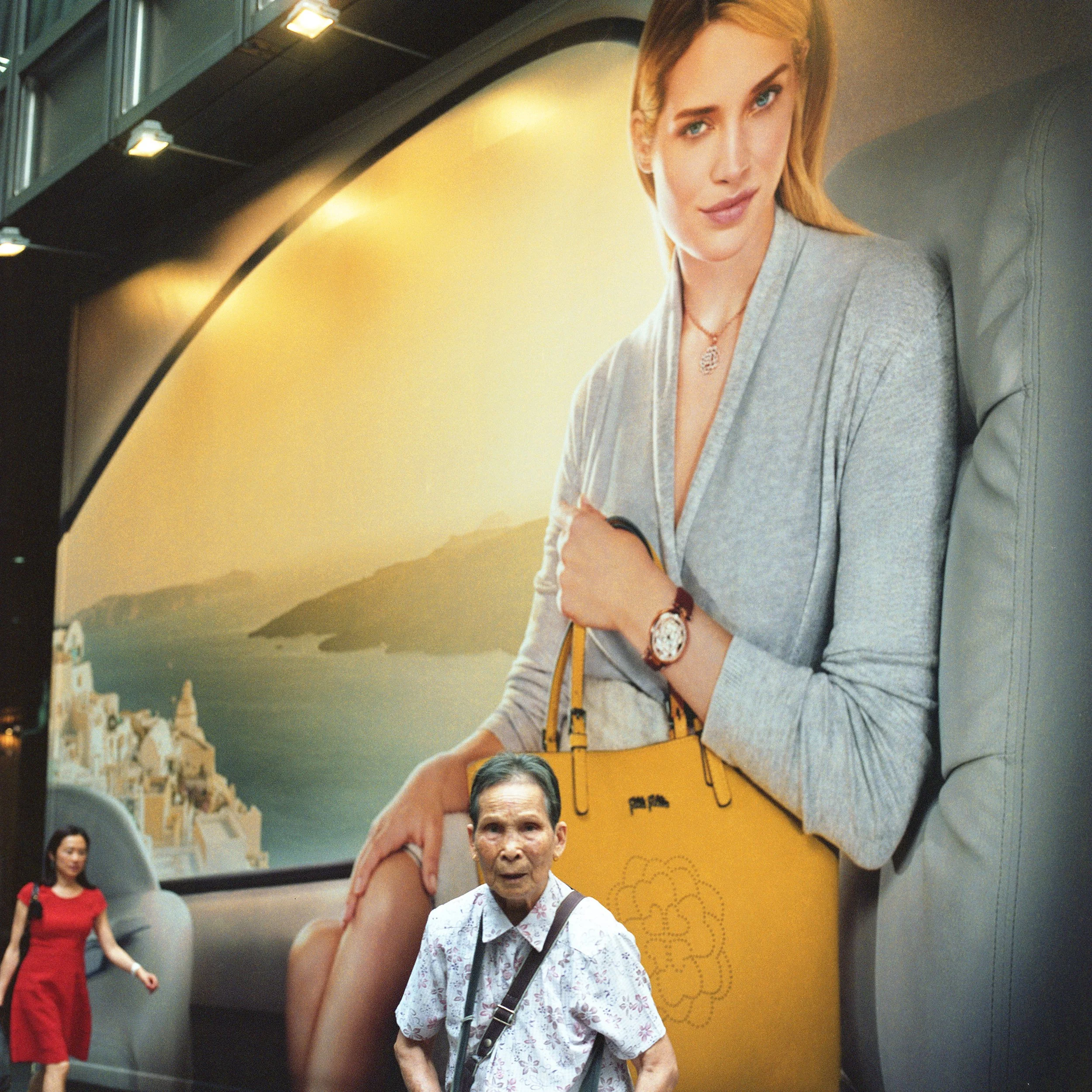 A large advertisement featuring a woman holding a yellow handbag while sitting on a gray sofa. The background shows a scenic view of water and landscape. In front of the advertisement, an elderly woman with short gray hair, wearing a floral blouse, s