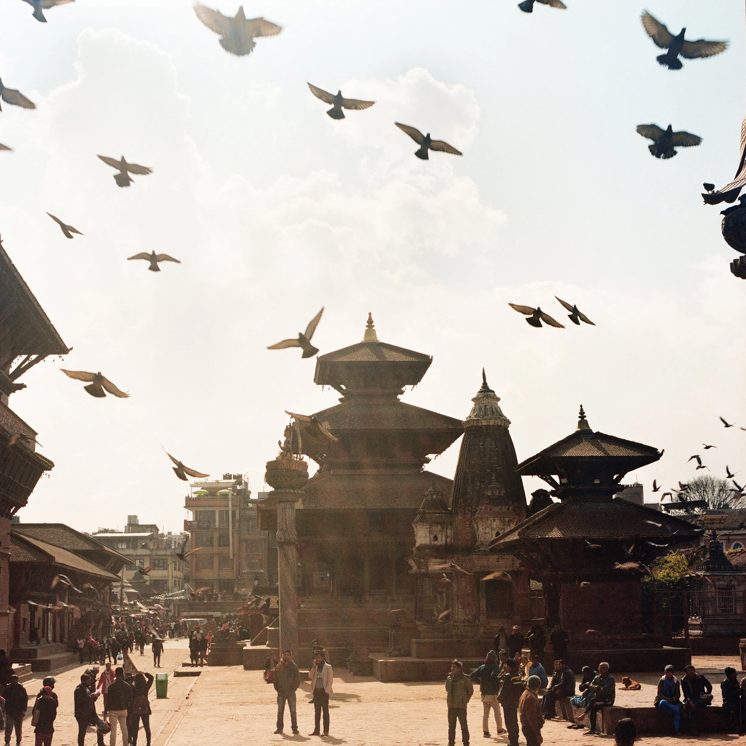 In Katmandu, People walking and sitting in a busy outdoor marketplace with traditional pagoda-style temples in the background, under a partly cloudy sky with birds flying overhead.