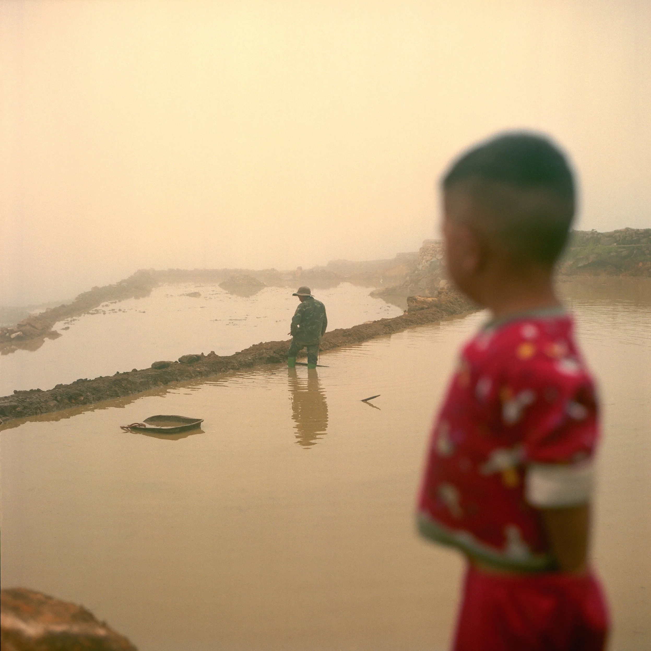 in Sa Pa, Vietnam, A foggy landscape with a young boy in the foreground wearing a red shirt with a pattern, looking at a man standing on a narrow mud path in a muddy body of water. 