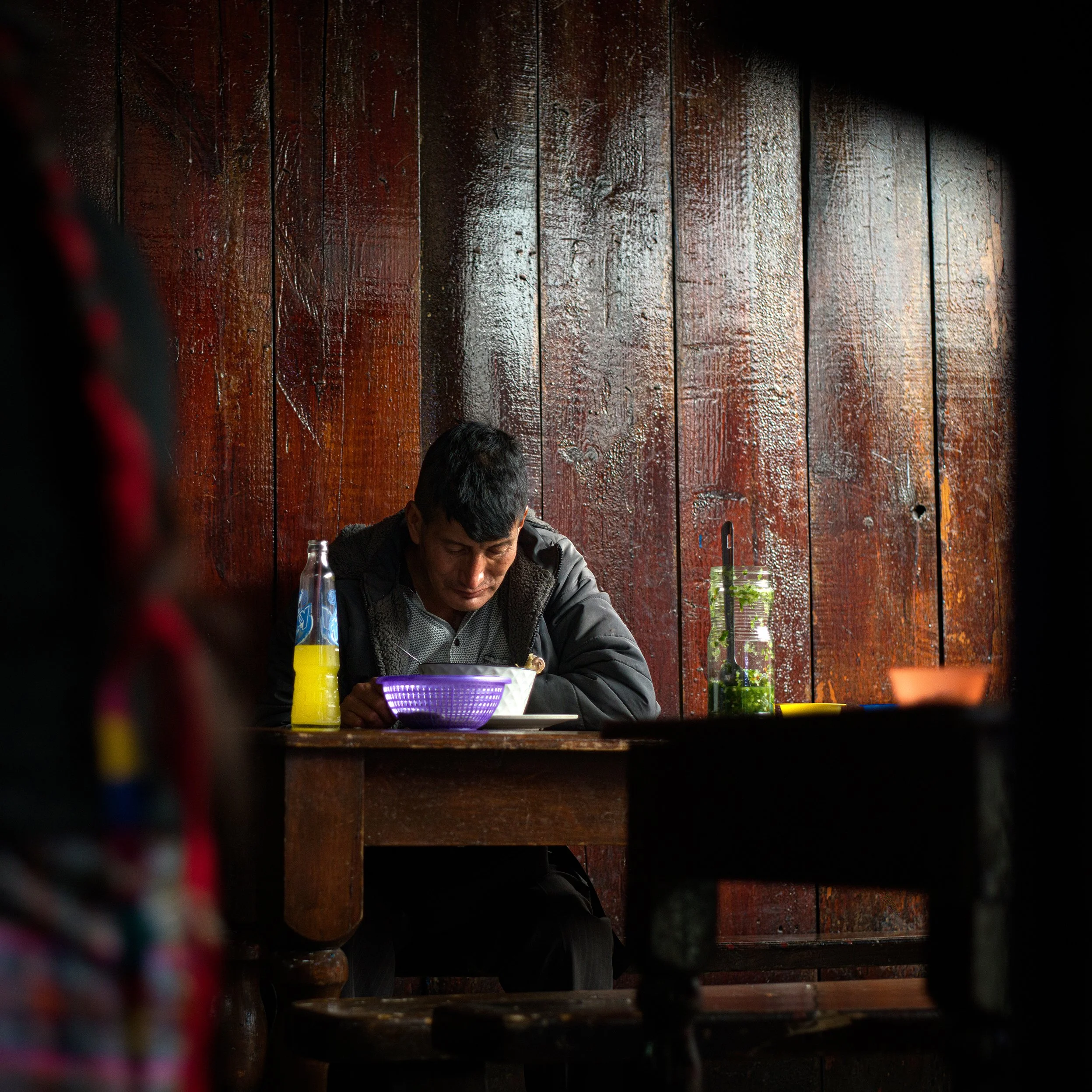 In Chichicastenango market, Guatemala, A man with dark hair, sitting at a wooden table against a dark, wet wooden wall, eating from a white bowl. A yellow drink in a glass bottle and a jar with green herbs are on the table.