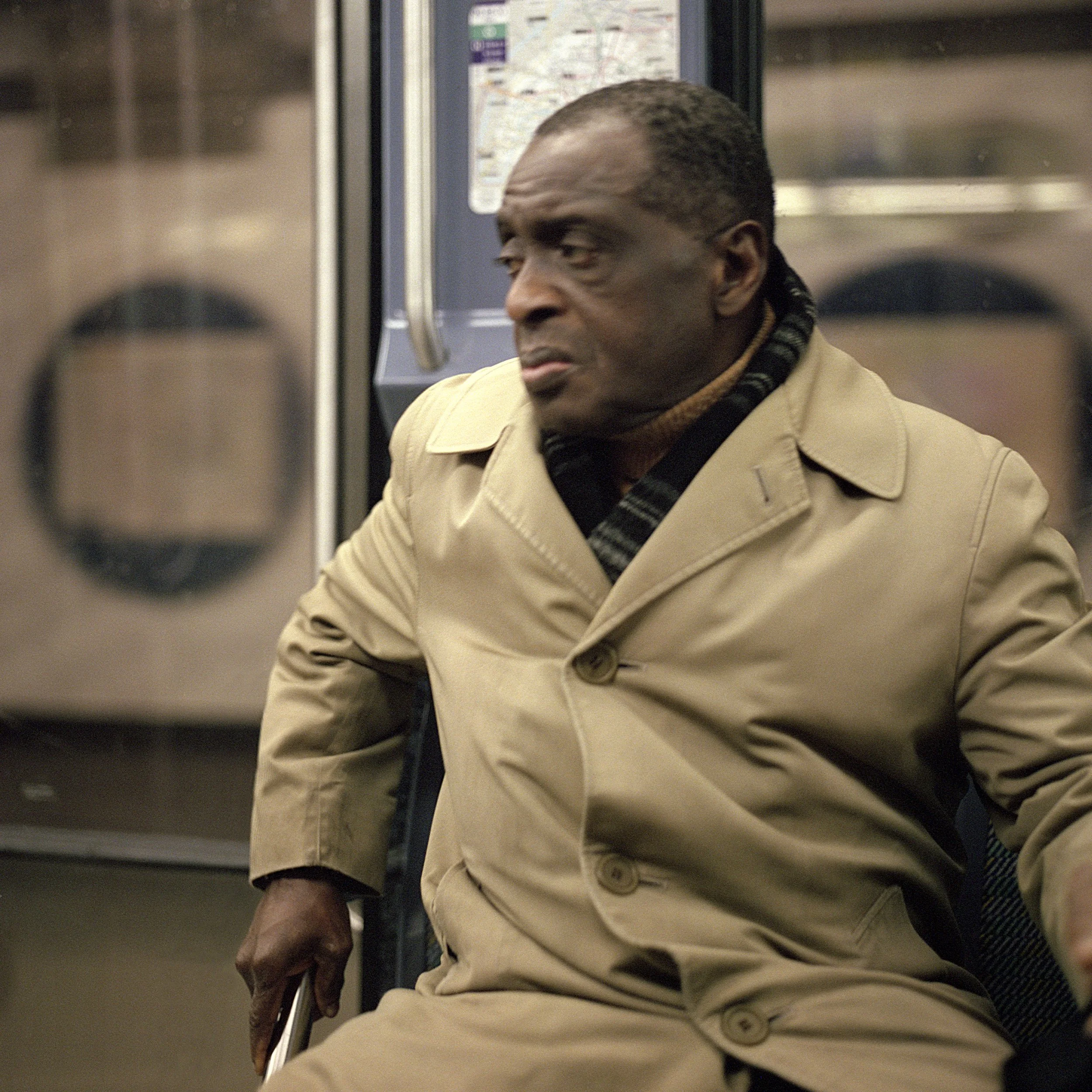 Man sitting on a in Paris Subway, wearing a tan trench coat and a scarf, looking serious.