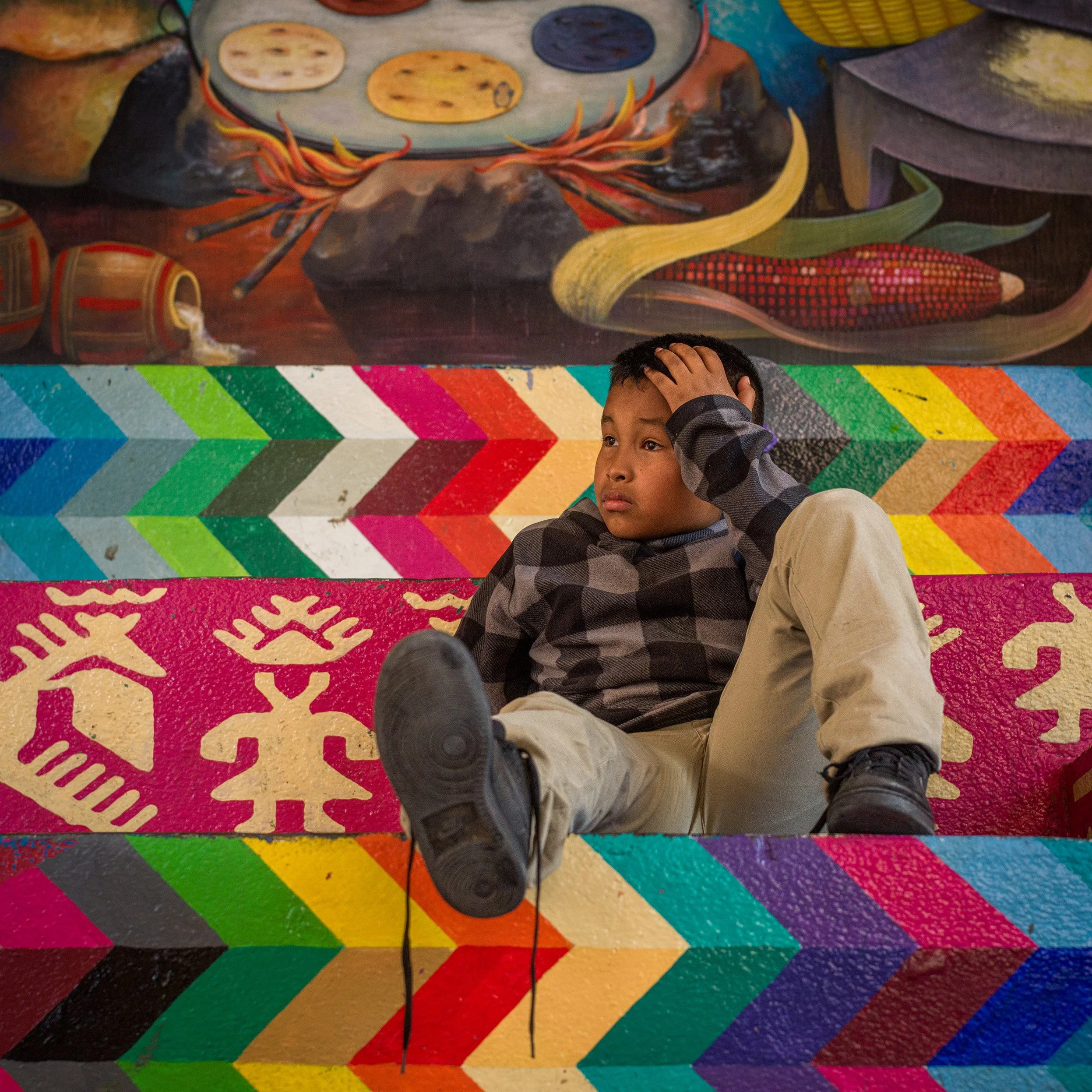 In Lake Atitlan, Guatemala, A Young boy sitting on colorful stairs with hand on his head, in front of a mural depicting traditional foods and utensils.