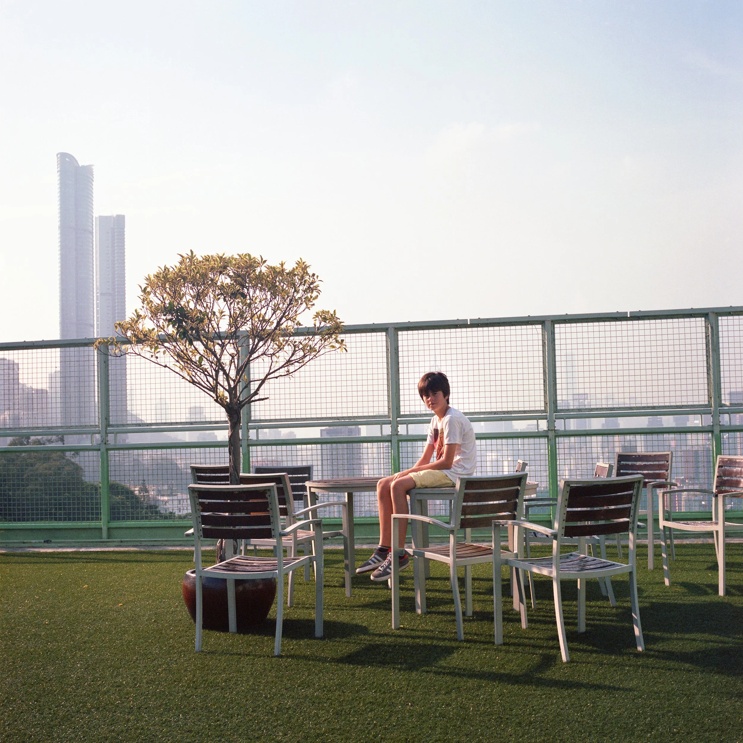 A young person sitting on a table on a rooftop terrace with city skyscrapers in the background, near a potted tree and chairs.