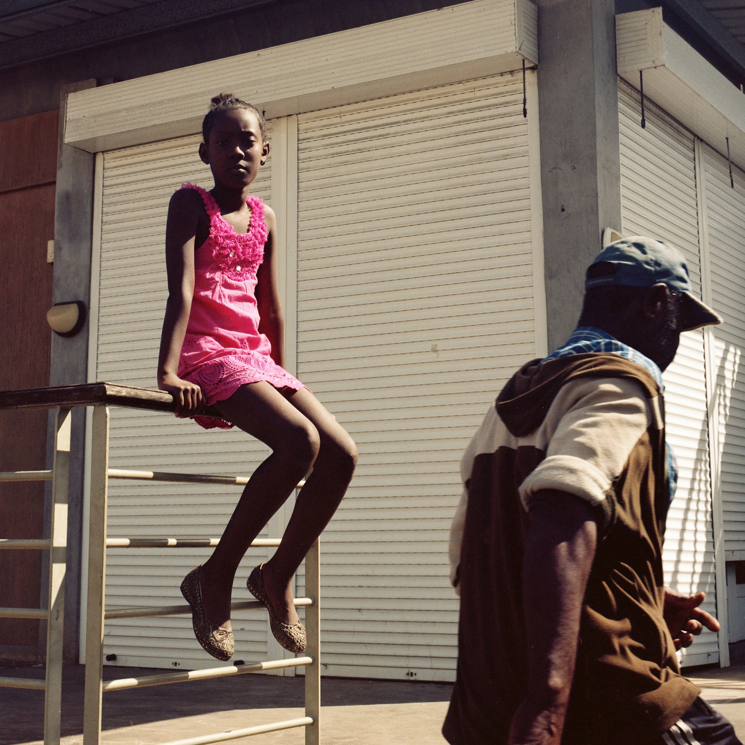 A young girl in a pink dress sitting on a metal railing, with an older man wearing a cap and jacket walking past in the background, outside a building with closed white shutters.