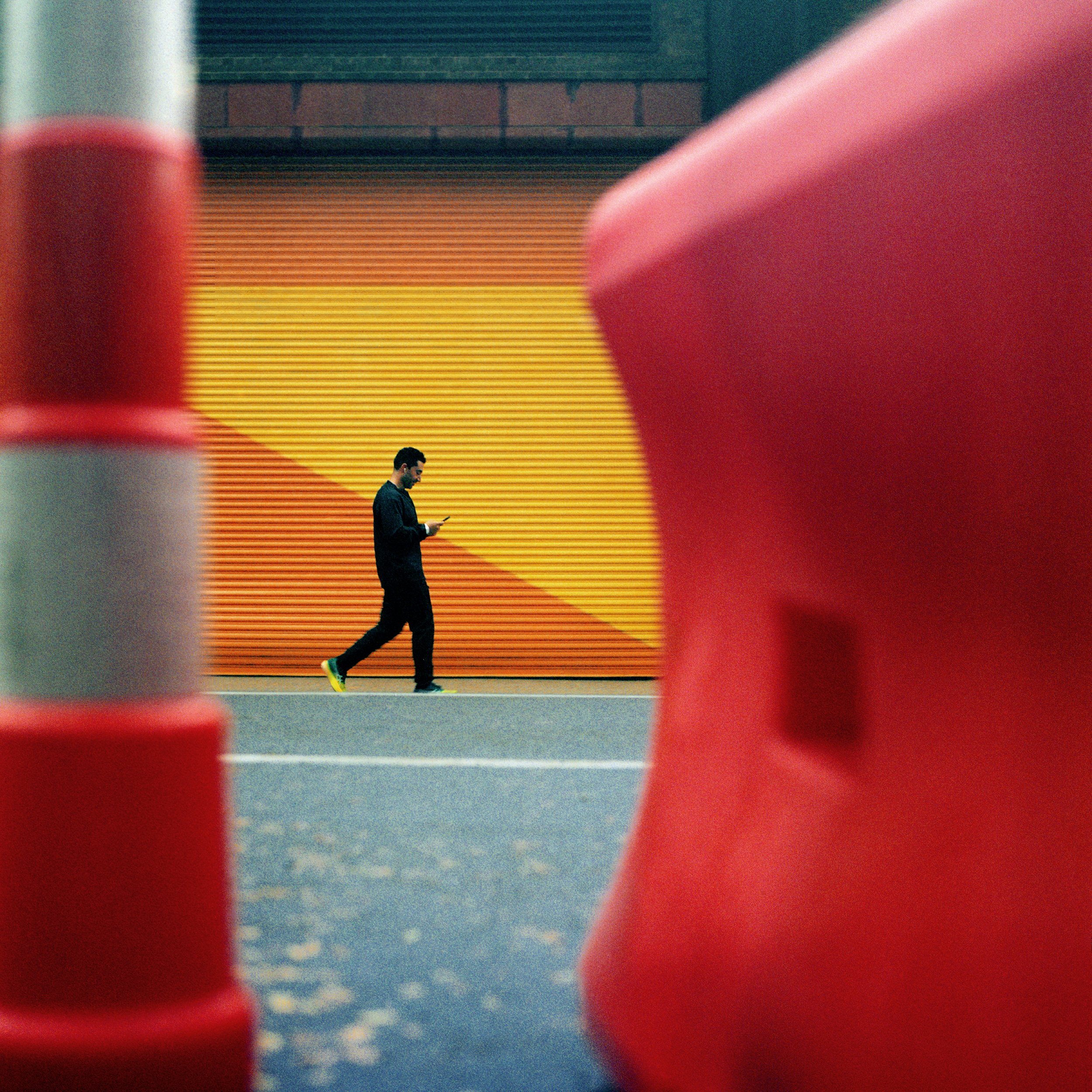 New York, A man in black clothing walking on a street, seen through two red objects in the foreground, with a colorful striped wall in the background.