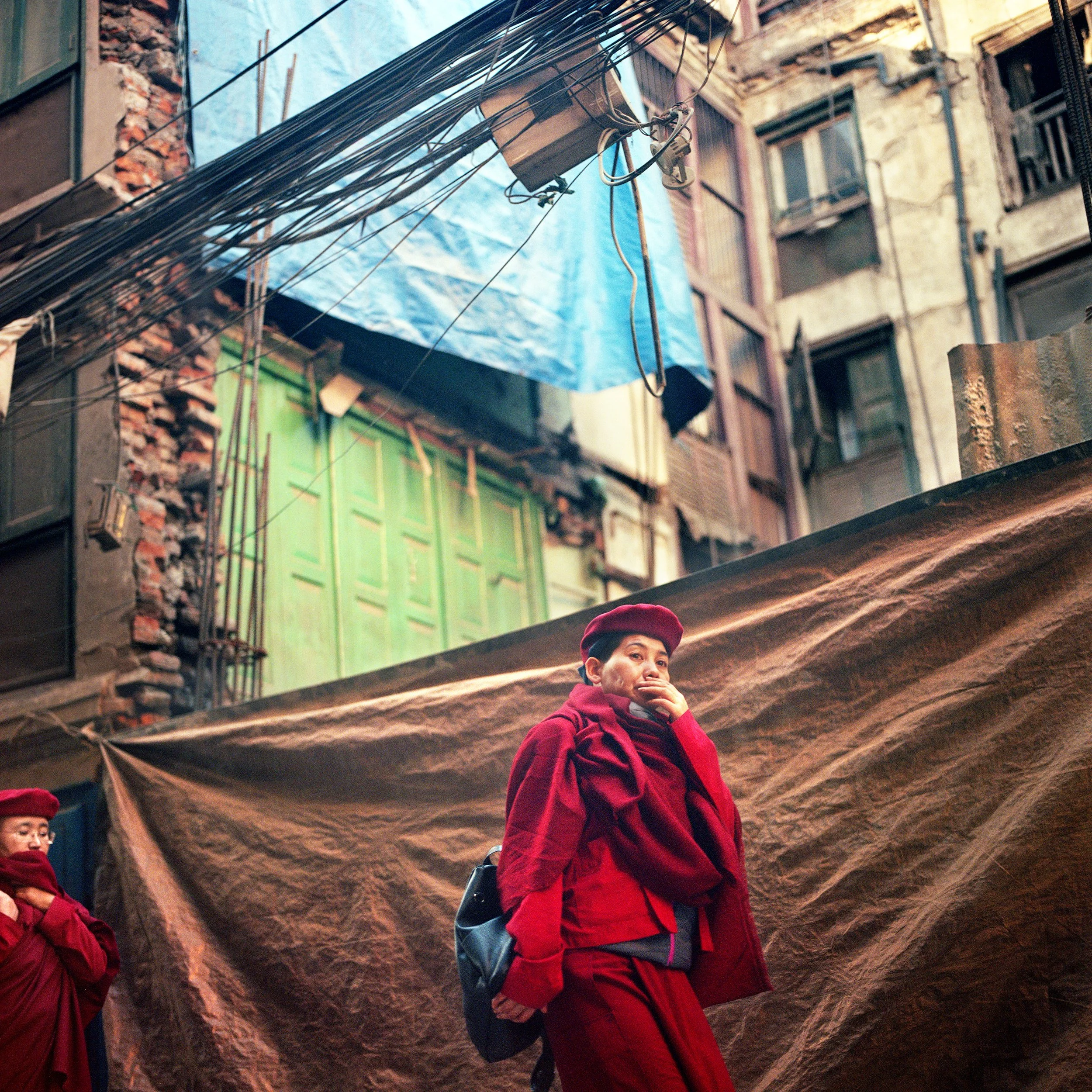A woman dressed in red standing in an outdoor urban alleyway with tall, worn buildings and tangled electrical wires overhead, covering her mouth with her hand.