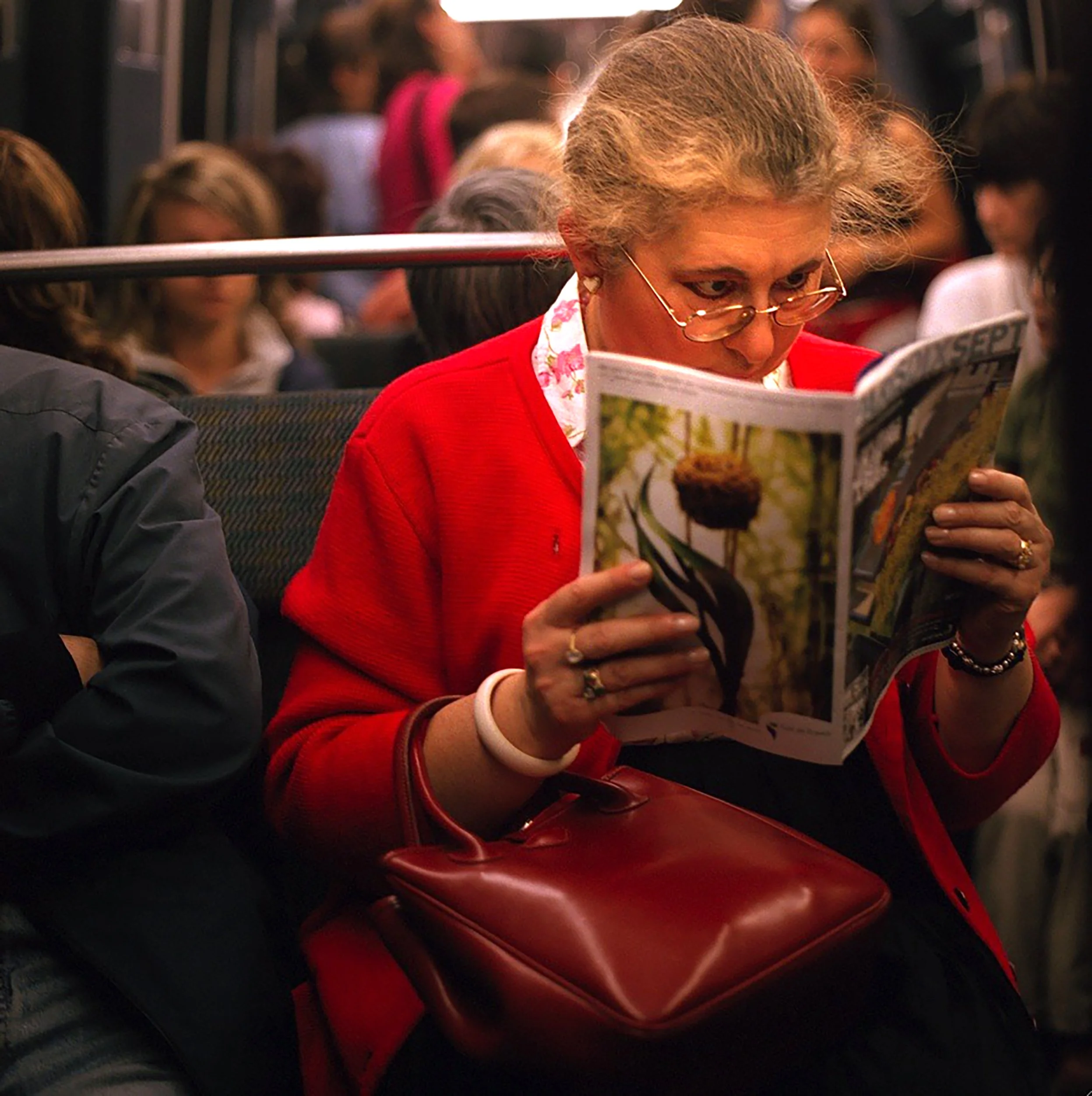 In Paris Subway, A woman with gray hair and glasses reading a magazine on a bus, with other passengers in the background.