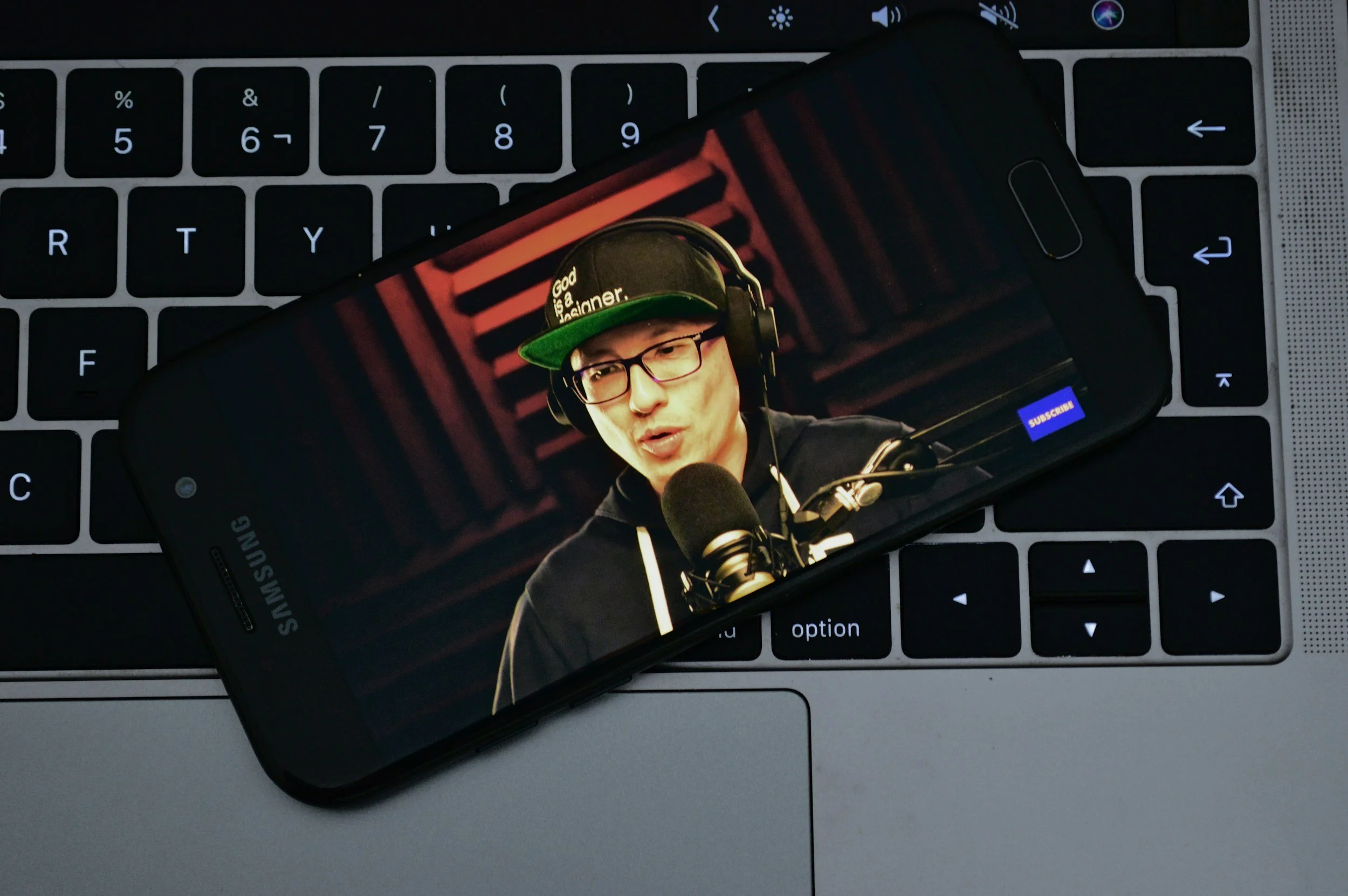 A smartphone lying on a laptop keyboard showing a man wearing glasses, a black hoodie, a black cap with green brim, and headphones, speaking into a microphone in a recording studio.