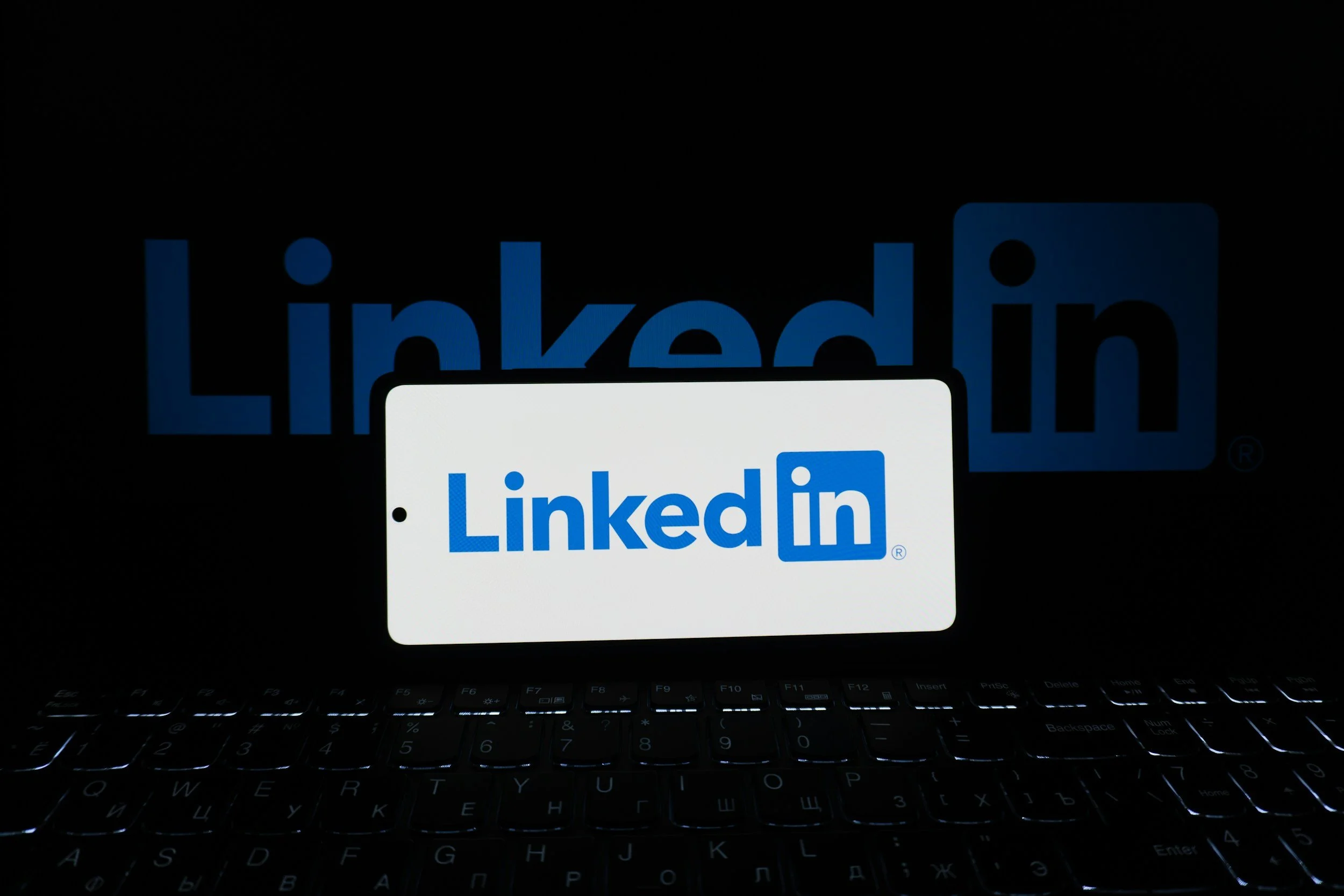 A computer screen displaying the LinkedIn logo in the background and a smartphone with the LinkedIn logo in the foreground, on a dark keyboard.