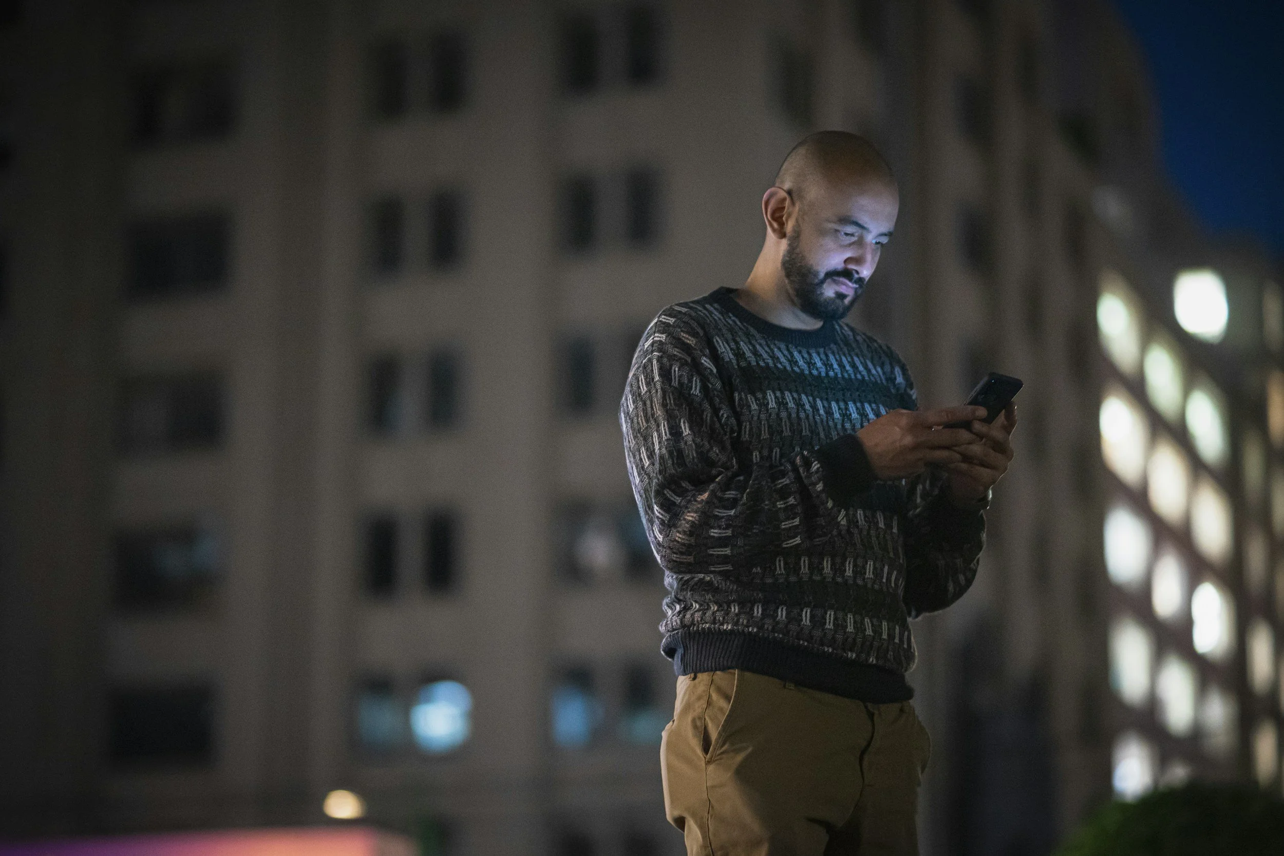 A man with a beard and a shaved head standing outside Dubai at night, looking at his phone with the glow illuminating his face. Behind him is an apartment building with lit windows.