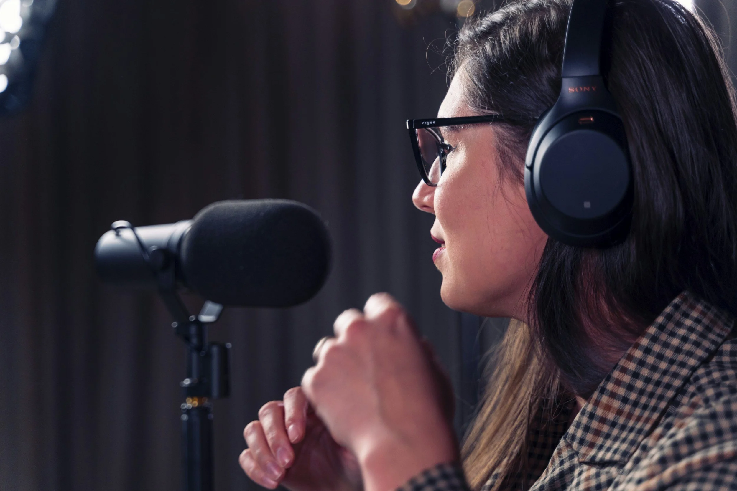 A woman speaking into a microphone, wearing large headphones and glasses, with long dark hair and a checked shirt.