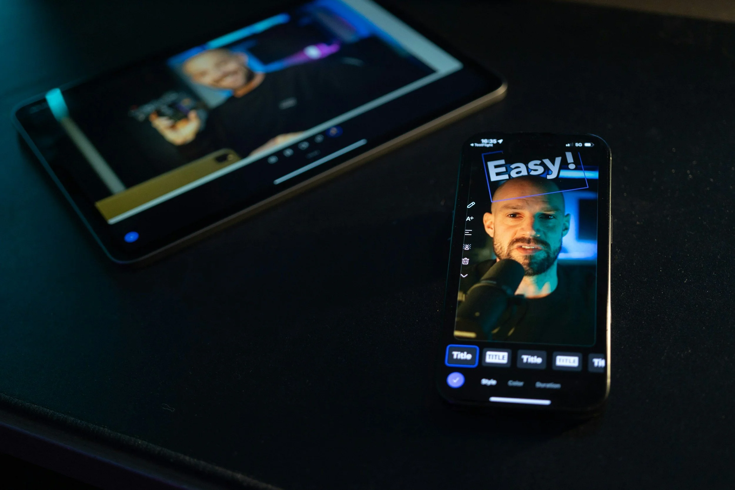 A smartphone on a dark surface displays a photo of a man speaking into a microphone with the word "Easy!" in large text above. A tablet in the background shows a different photo of the same man, slightly out of focus.