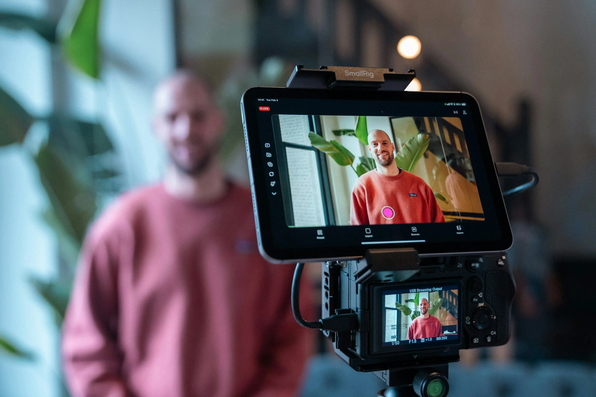 A man in a red sweater being recorded on a podcast camera in Dubai, with the camera's viewfinder showing his smiling face and background of green plants and a window.