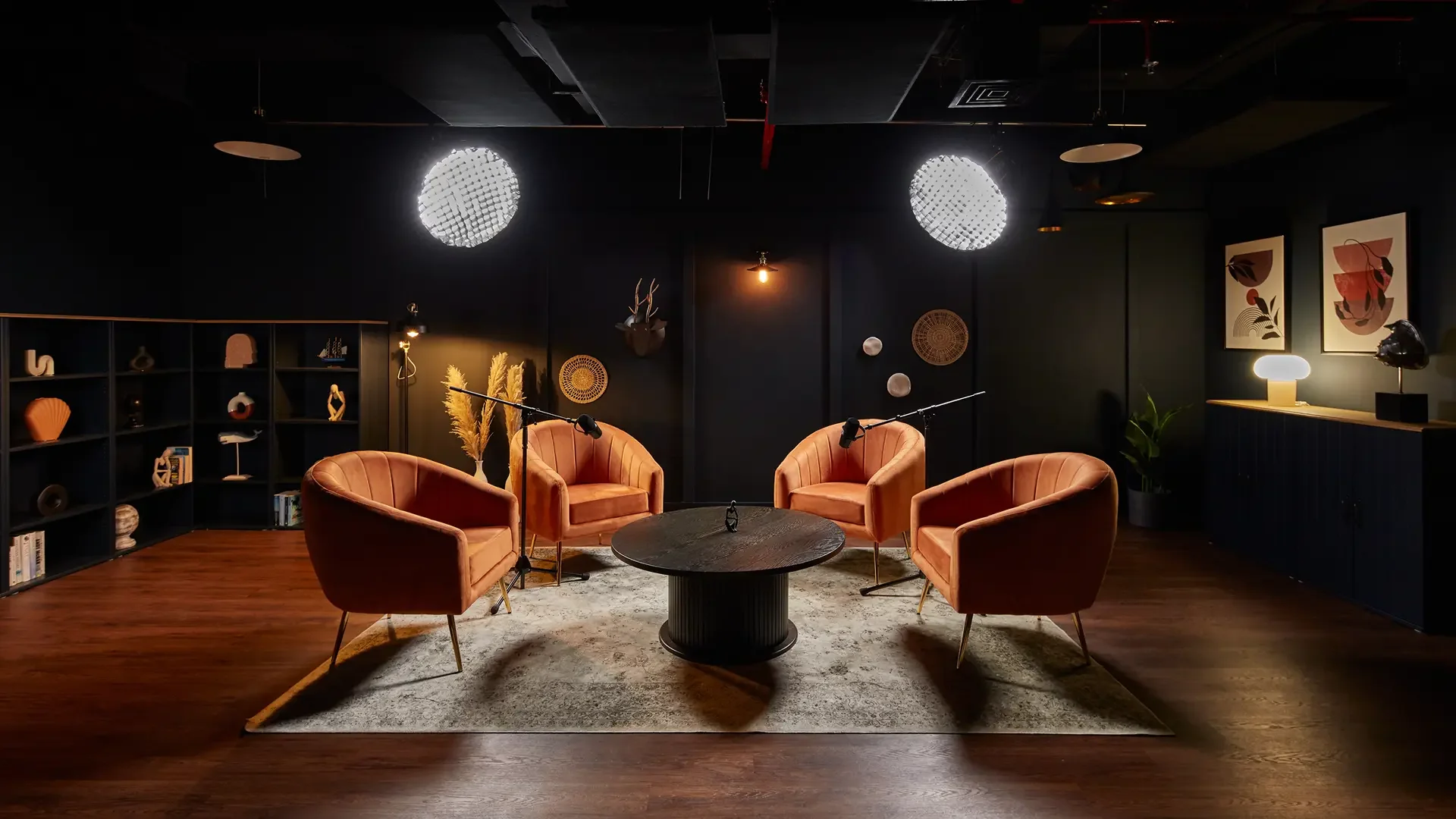 A set of four peach-colored velvet armchairs arranged in a semicircle around a round black coffee table in a dimly lit modern room, with black walls, wooden floor, and decorative items on the shelves and walls.