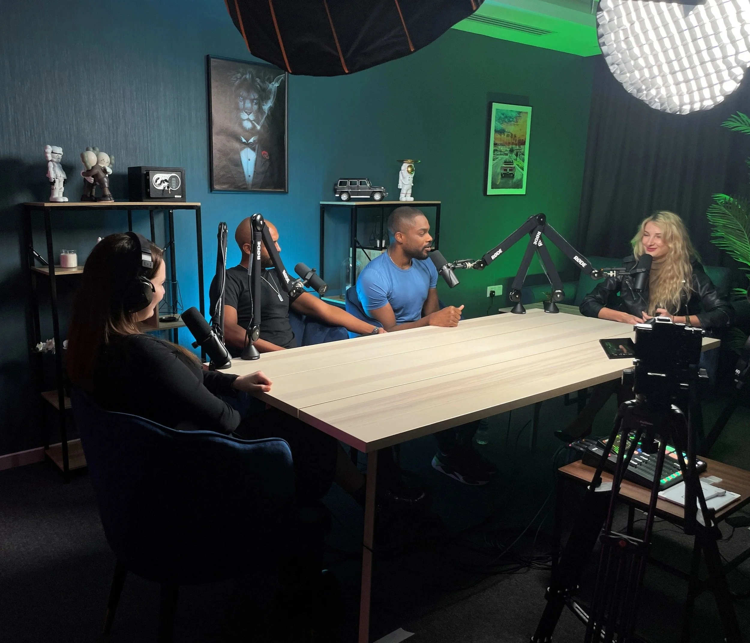 A podcast recording in Dubai with four people sitting at a long table with microphones, headphones, and a camera in front of the table. The room has dark walls, colorful artwork, and decorative items on shelves.
