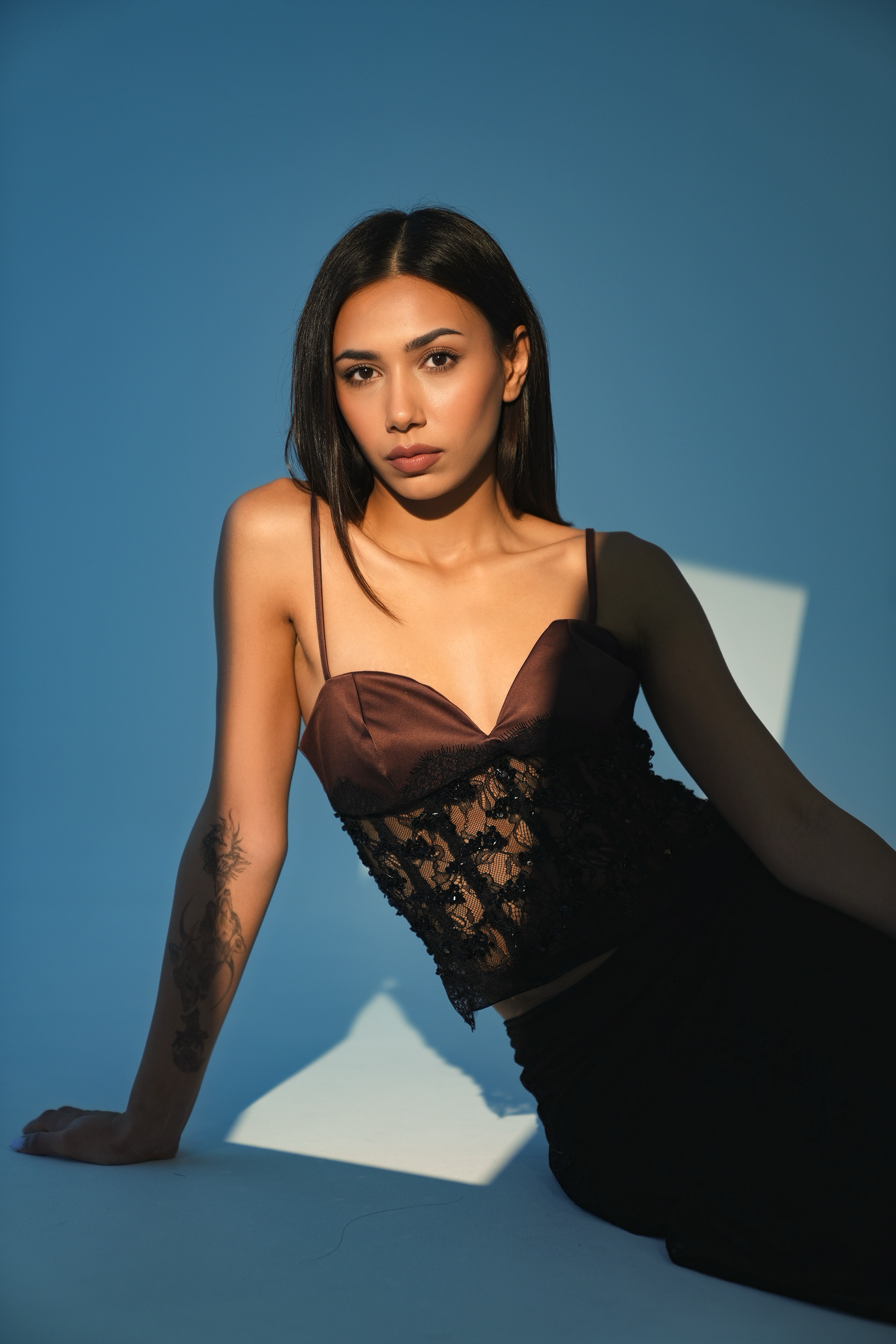 A woman with long black hair and a tattoo on her left arm, wearing a dark brown and black lace dress, sitting on the ground with a blue background and a shadow of a square behind her.