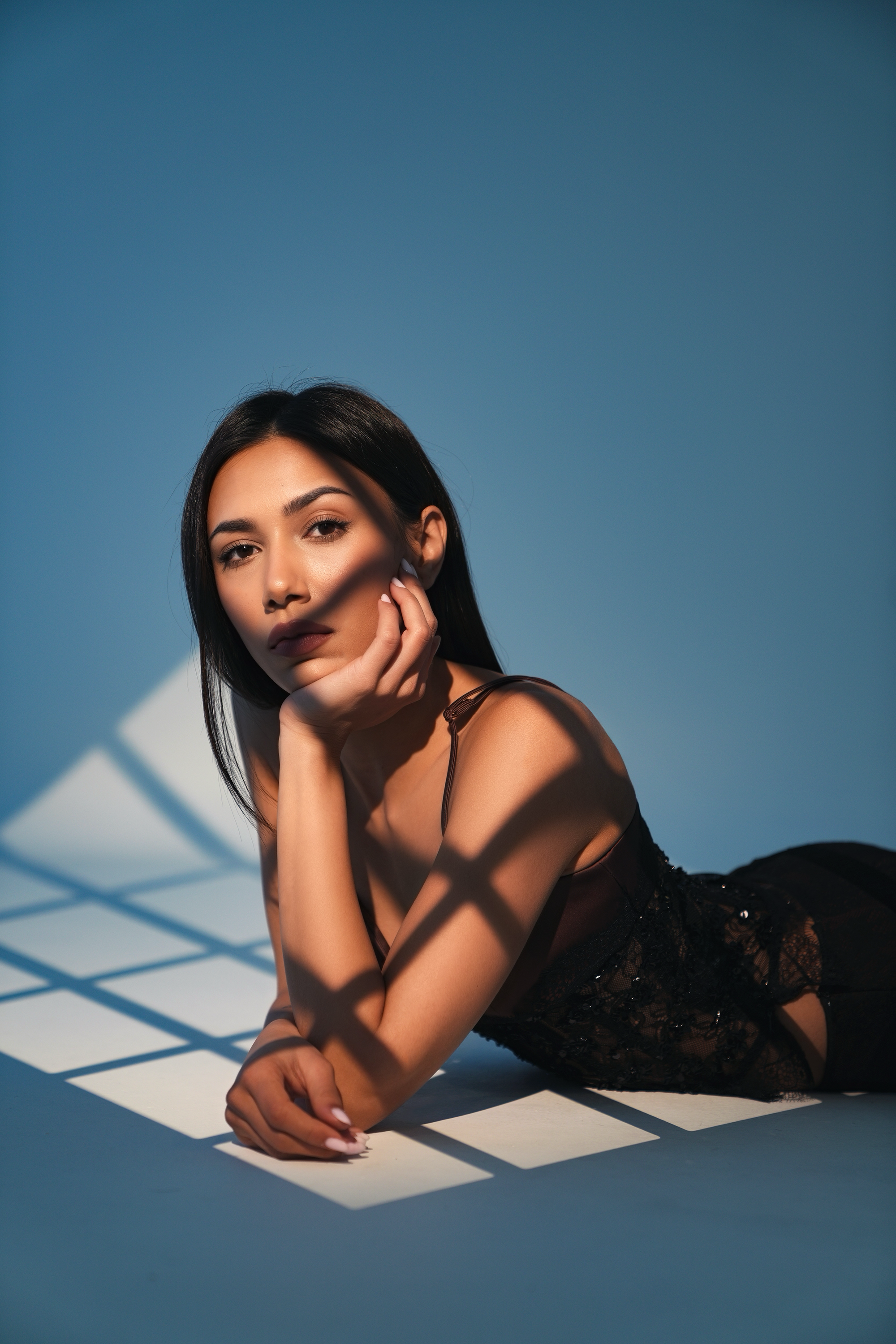 A woman with dark hair and makeup lying on the floor, resting her head on her hand, with shadows of a grid pattern cast on her and the background.