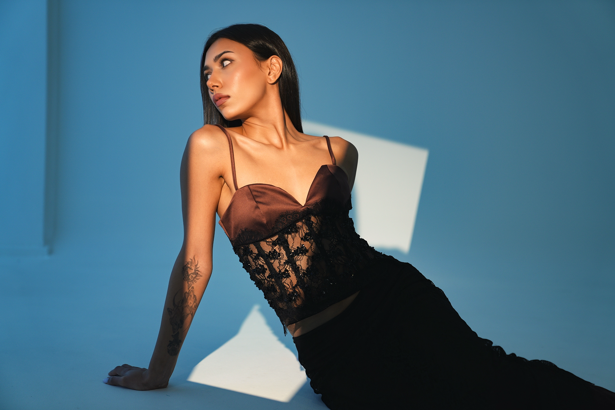 A woman with long dark hair, wearing a brown and black lace dress, is sitting on the floor with her left hand resting on the ground, looking to her left against a blue background with a white rectangular shadow.