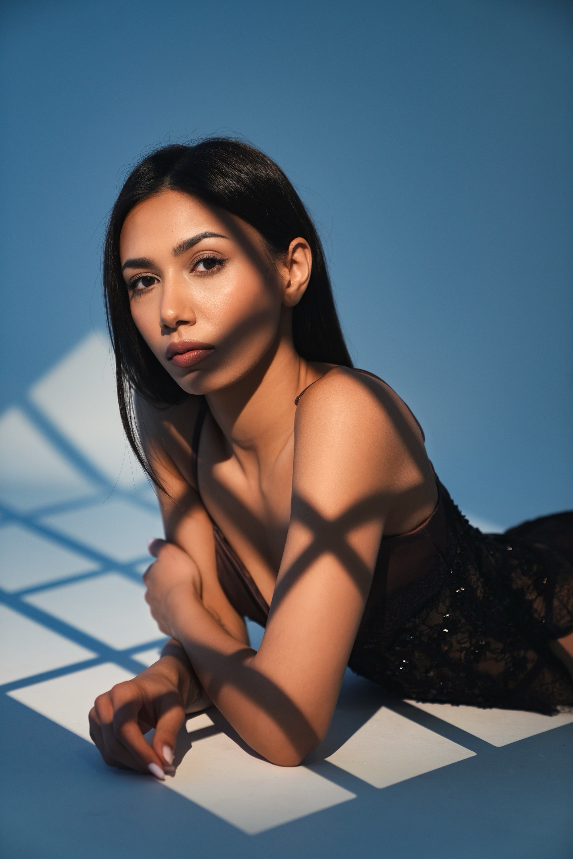 A young woman with black hair and wearing black lace clothing, lying on her side on a white floor with shadows cast by a grid pattern, against a blue background.