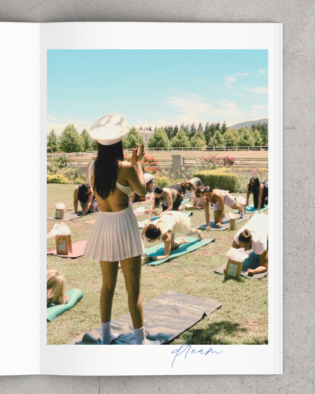 A group of women participating in an outdoor yoga class on a sunny day, with a woman in a white hat and pleated skirt standing in front, leading the class. The women are on yoga mats on the grass, doing poses. There are trees and a fence in the backg