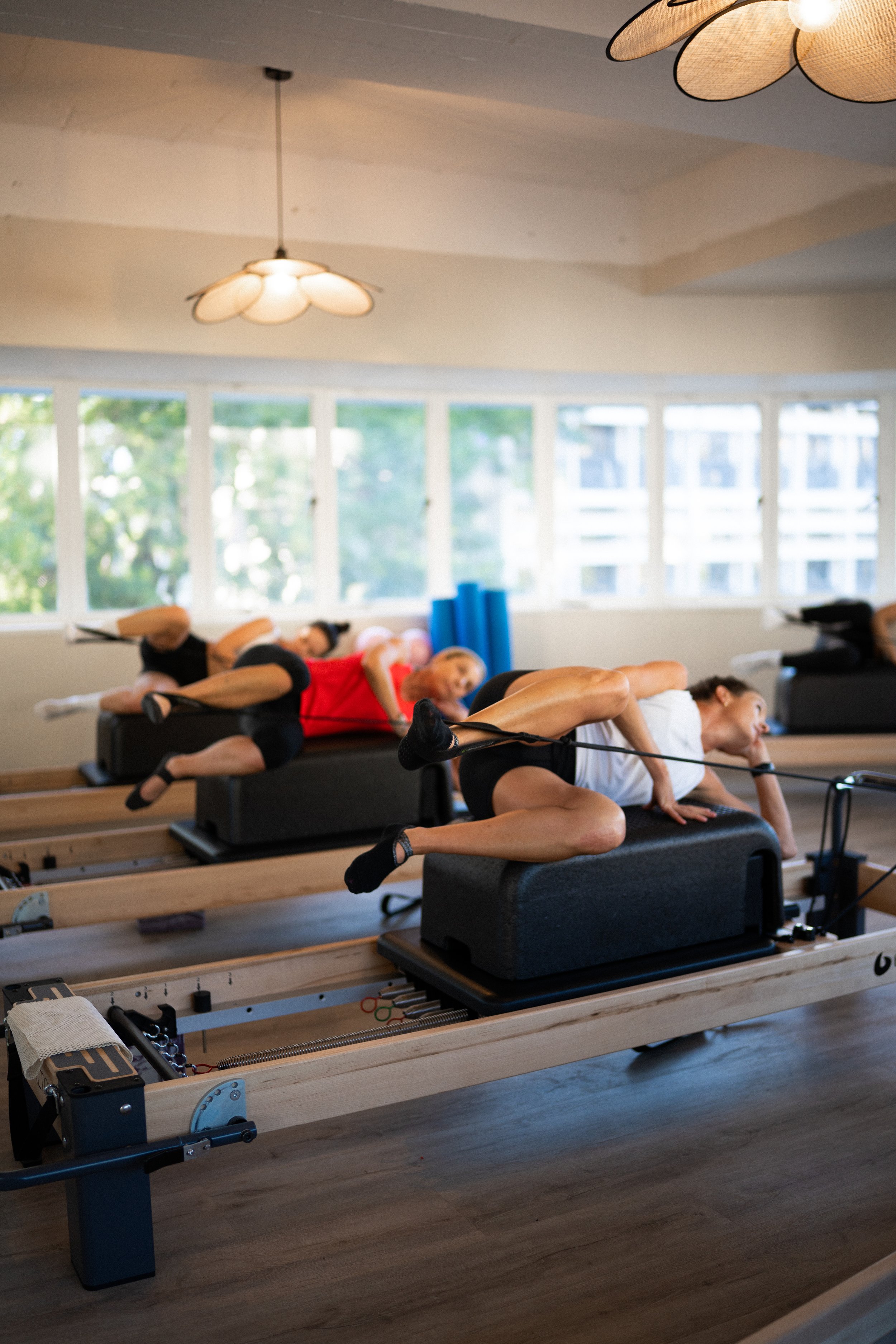 People participating in a Pilates class on reformer machines in a bright studio with large windows.
