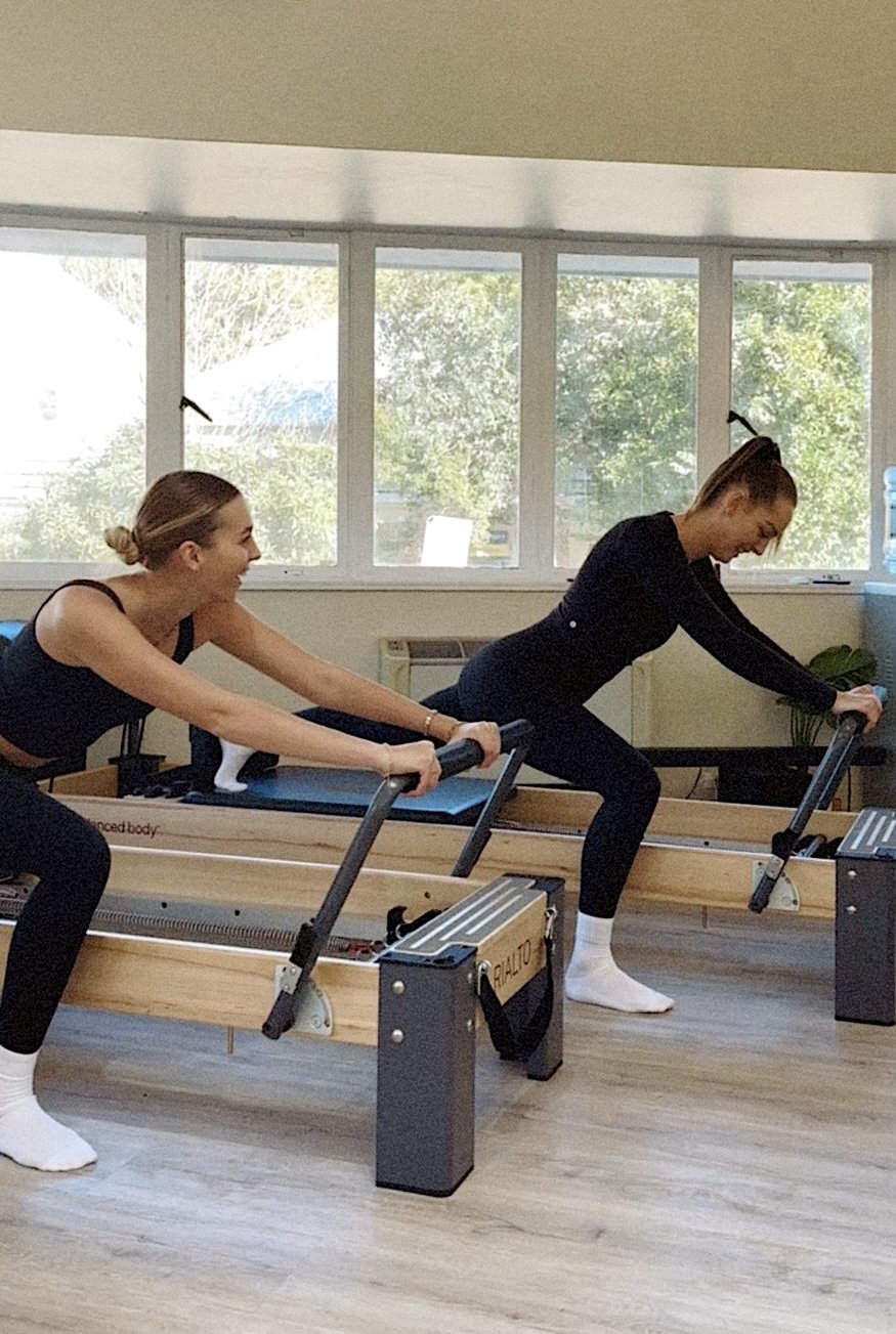 Two women using a reformer Pilates machine in a fitness studio, smiling and laughing.