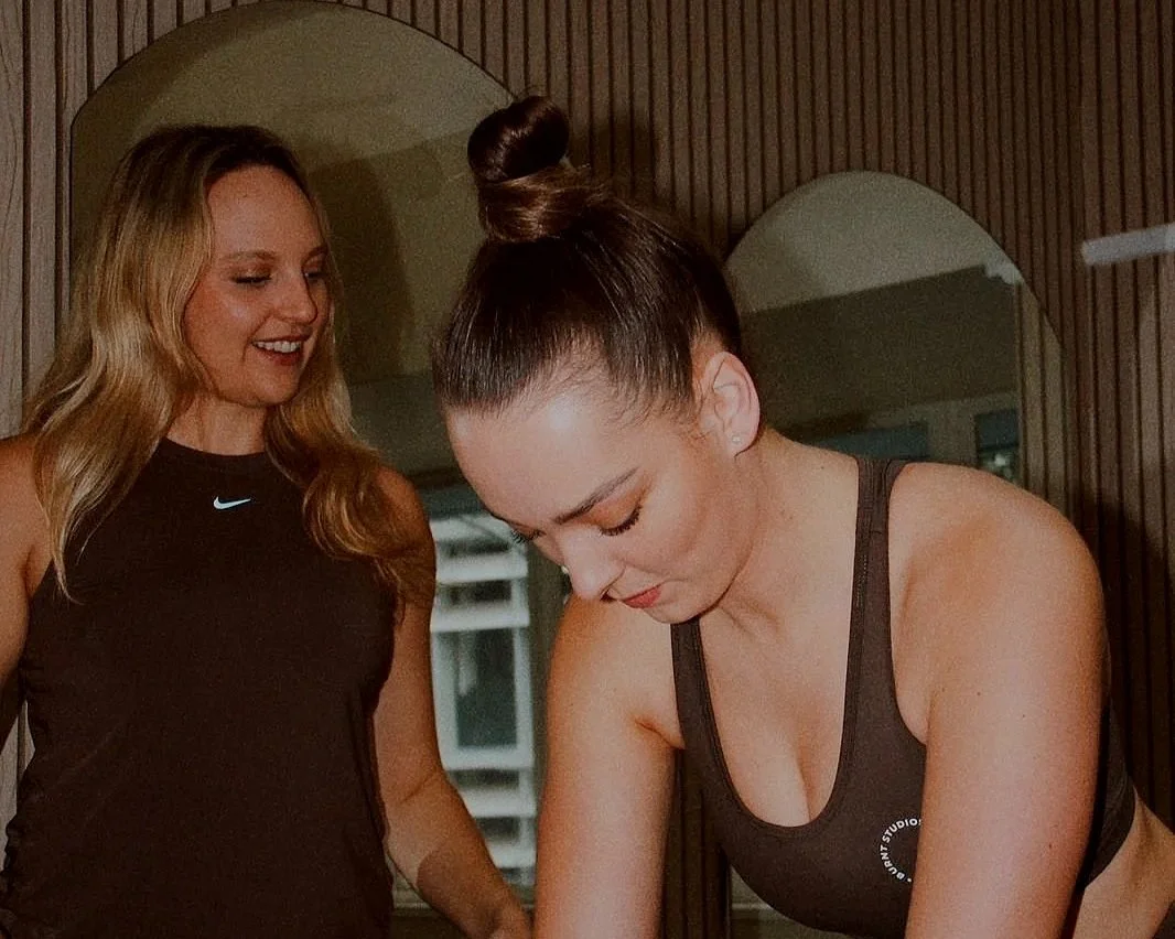 Two women in workout attire standing indoors near mirrors, one looking down and the other smiling.