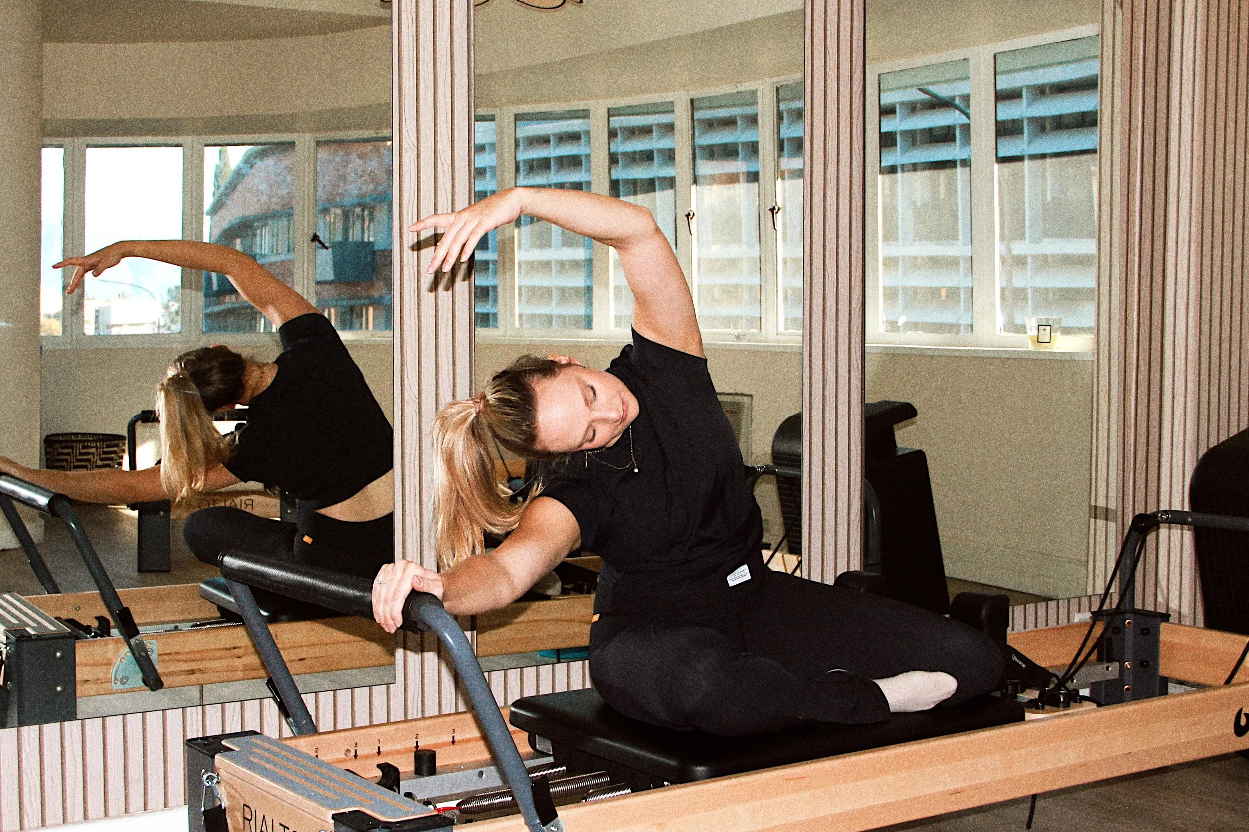 Woman on Pilates reformer machine, stretching with a mirror behind her reflecting her pose in a fitness studio with large windows.
