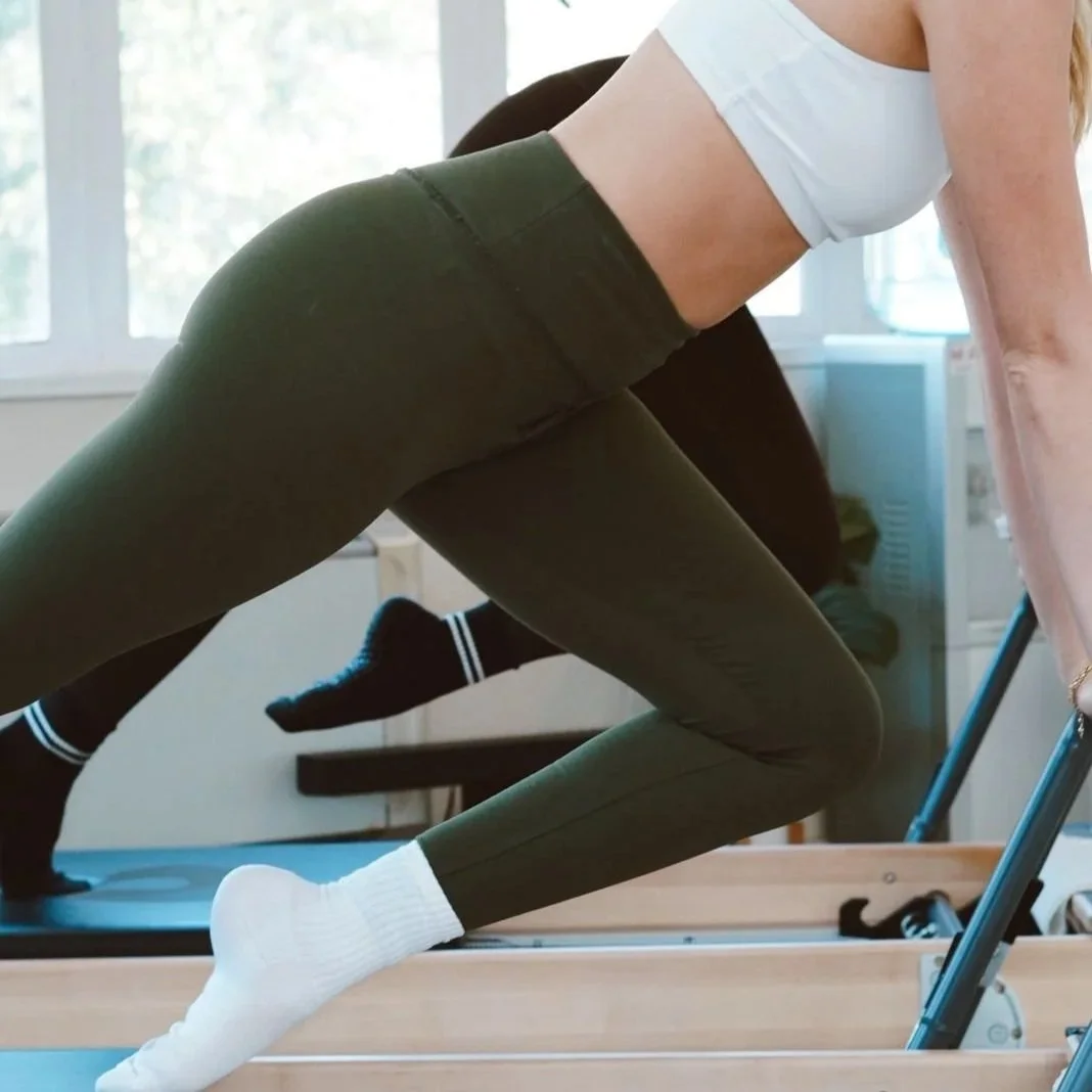 Person stretching on a wooden exercise machine, wearing dark pants, a white crop top, white socks, and black socks with white stripes on the treadmill, with sunlight streaming through windows in a bright room.