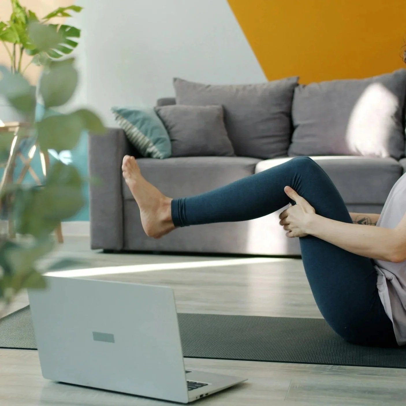 A person practicing abdominal exercises at home, with their legs raised and hands on their thigh, on a yoga mat in a living room with a gray sofa and green plants.