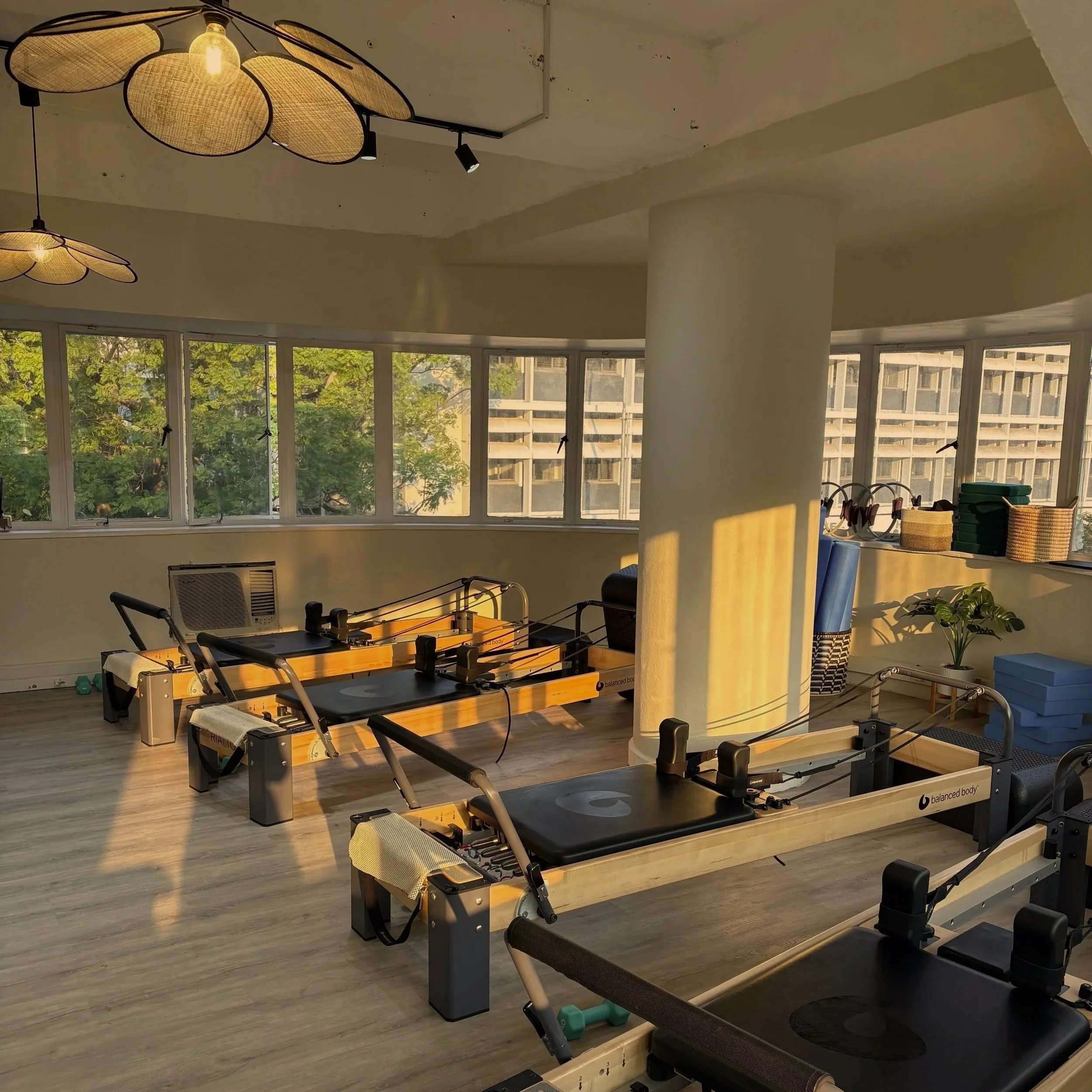 Pilates reformer machines in a bright, sunlit fitness studio with large windows, hanging lights, and fitness accessories