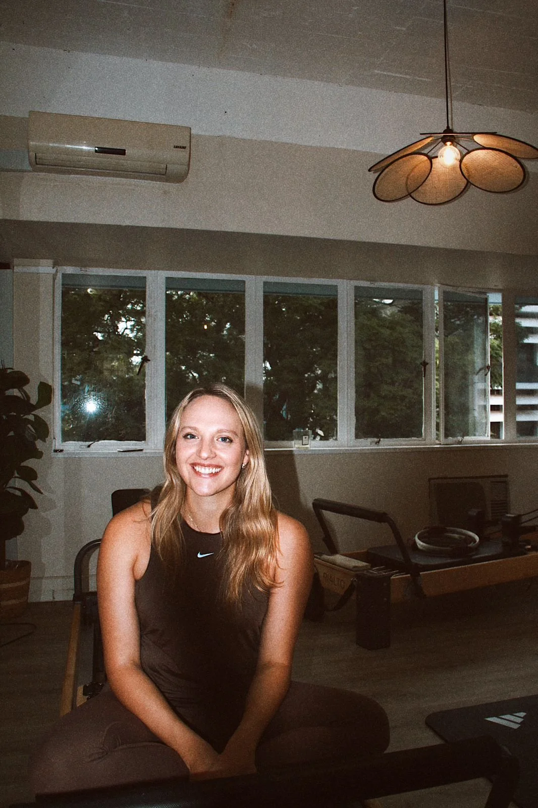 A young woman with long blonde hair, wearing a black sleeveless top, smiling and sitting on a chair in a room with large windows and indoor plants.